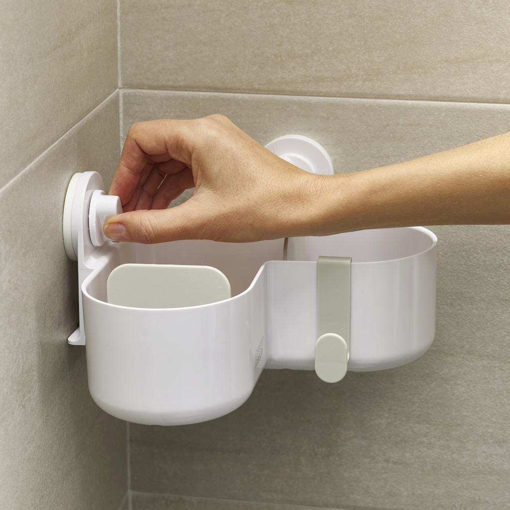 Una mano ajusta una cesta de ducha de plástico blanco montada en una pared alicatada. La cesta tiene compartimentos y un gancho.