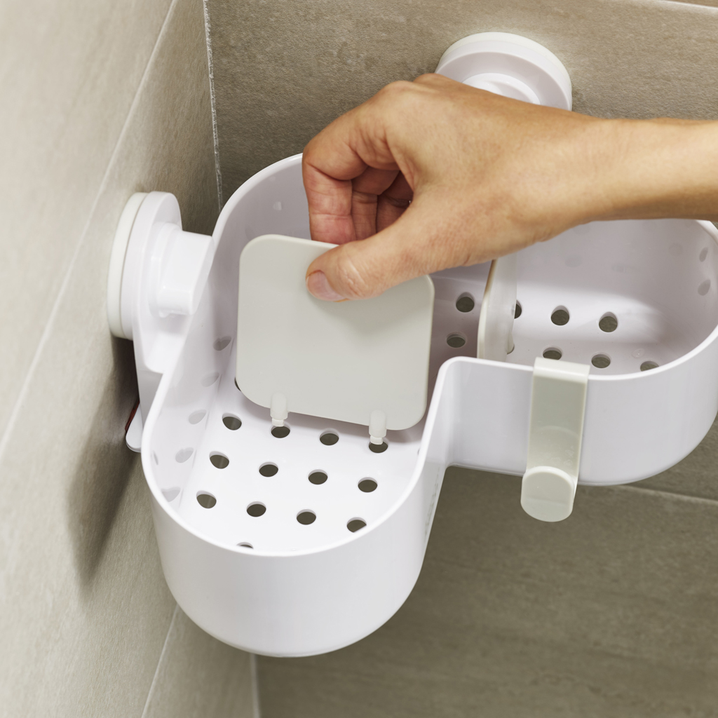 Una mano coloca una pieza cuadrada gris en una cesta de ducha de plástico blanco montada en una pared alicatada.