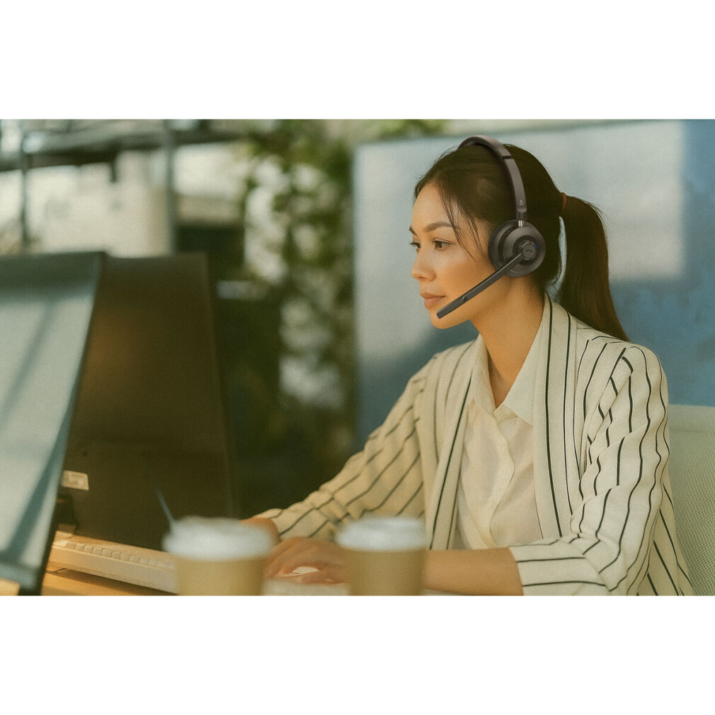Una donna con le cuffie lavora al computer. Si trova in un ambiente d'ufficio, con tazzine di caffè sulla scrivania. Indossa una giacca a righe.