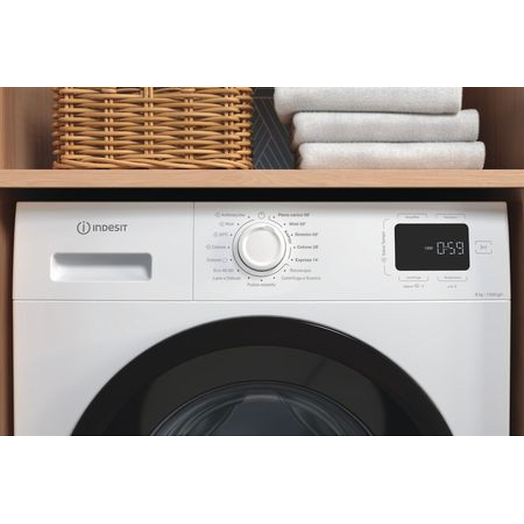 Una lavatrice Indesit bianca è sotto uno scaffale con un cestino di vimini e asciugamani bianchi piegati. Il display mostra '0:59'.