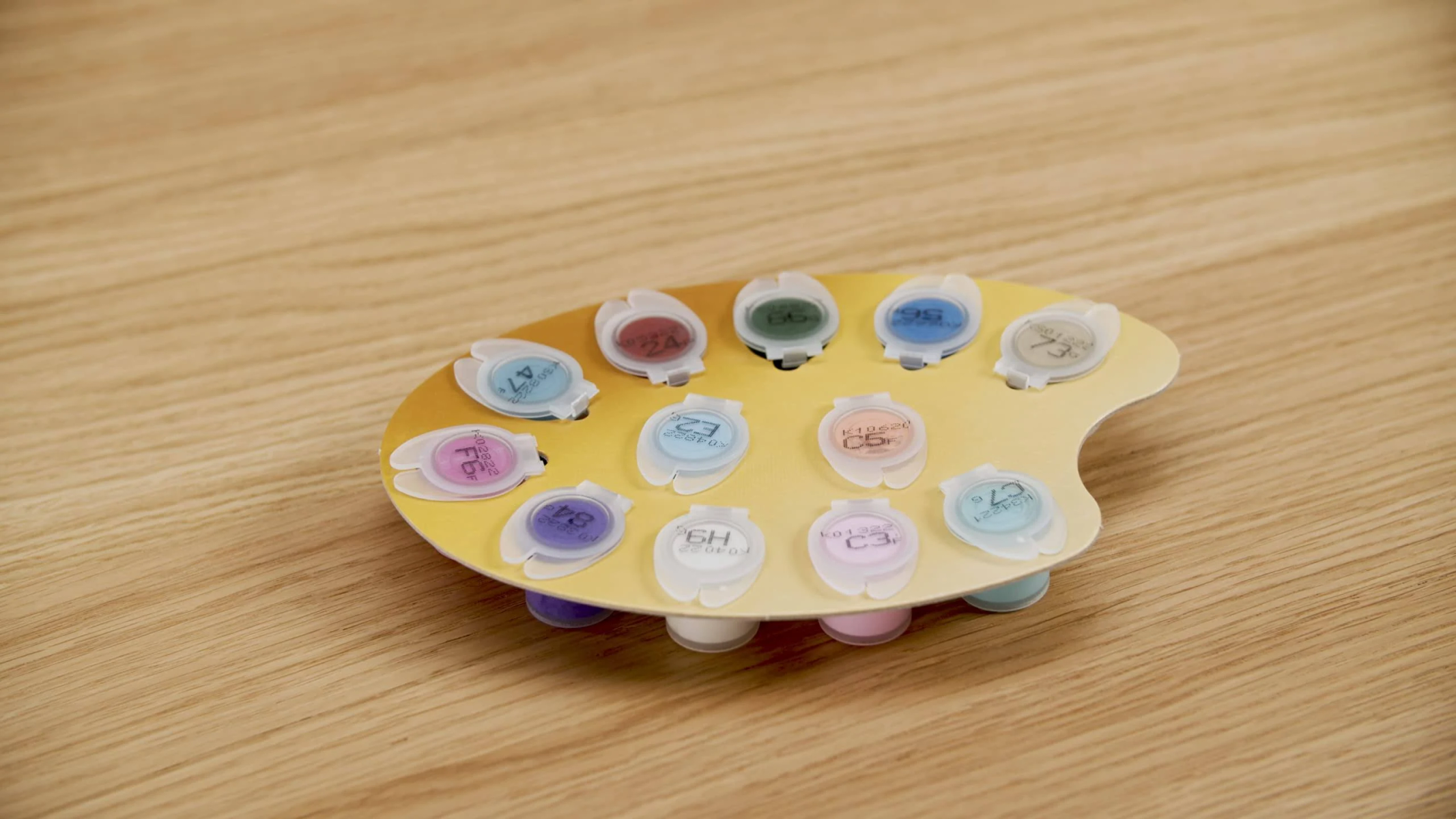 Eine gelbe Palette mit zwölf kleinen Farbtuben mit farbigen Deckeln auf einem Holztisch. Zahlen und Buchstaben stehen auf den Deckeln.