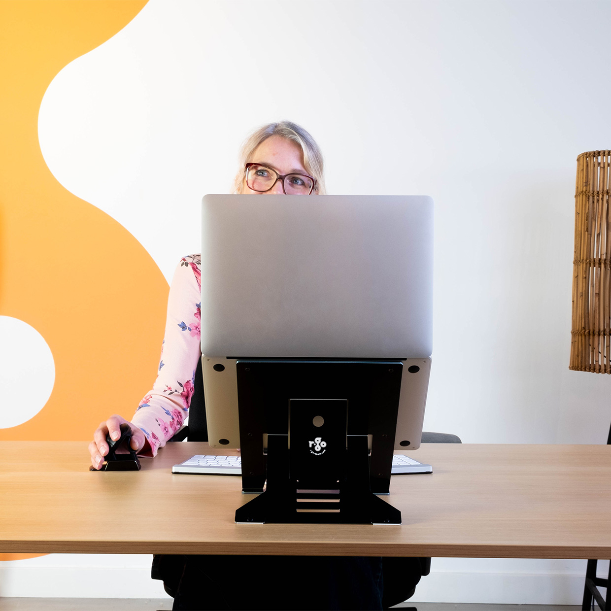 Frau mit Brille sitzt an einem Schreibtisch und benutzt einen Laptop. Vorne steht ein schwarzer Laptopständer. Der Hintergrund hat ein orange-weißes Design.