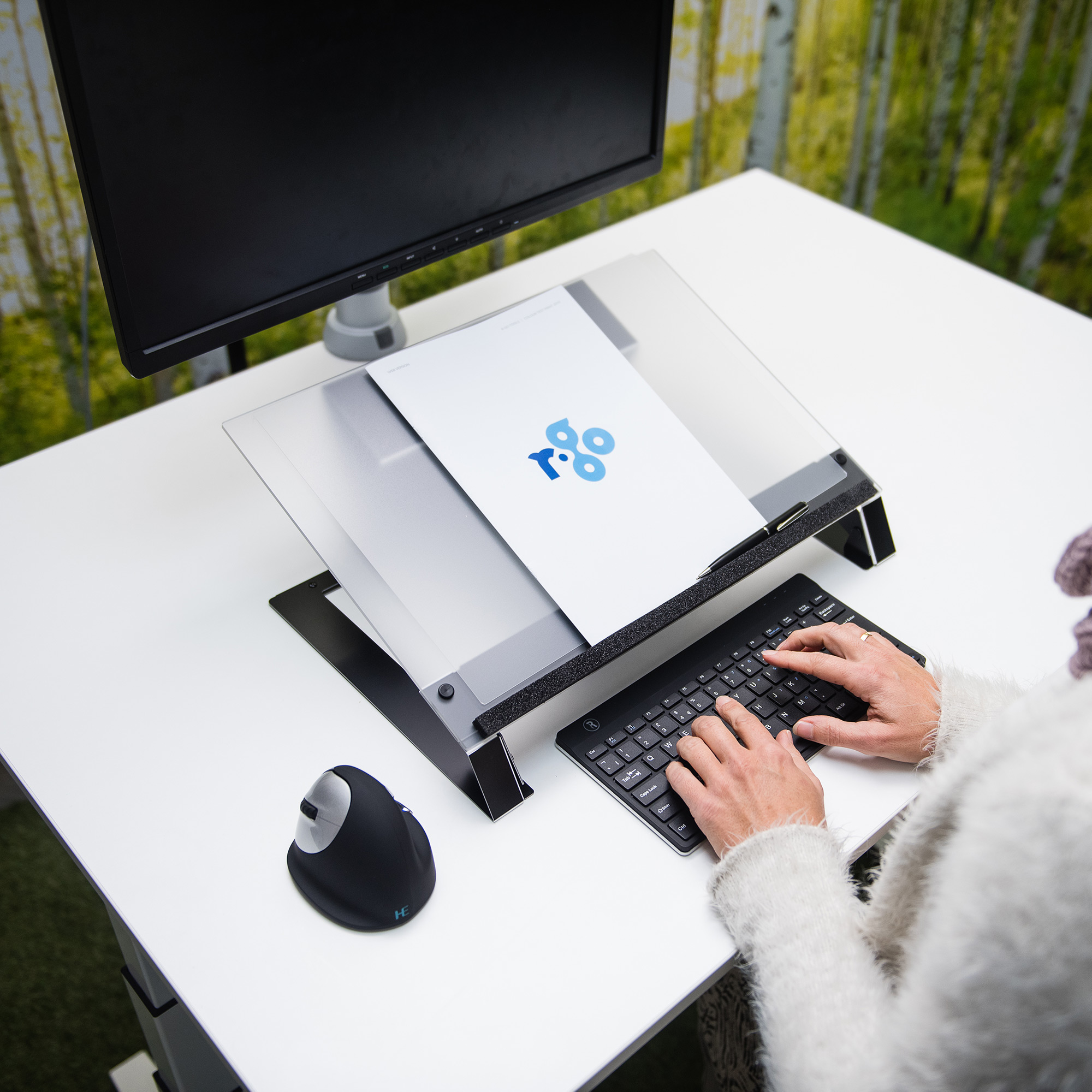 Eine Person tippt auf einer Tastatur an einem weißen Schreibtisch. Ein Monitor, Papier und eine Maus sind sichtbar. Grün-weißer Hintergrund.