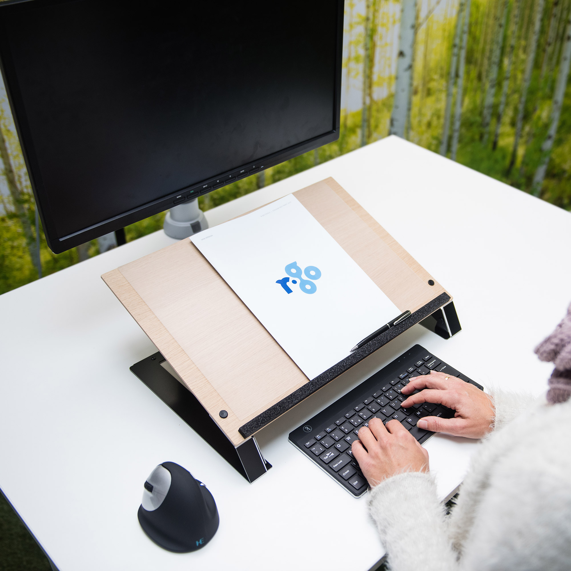Ein Schreibtisch mit Monitor, Tastatur, Dokument auf einem Ständer und einer Maus. Hände, die auf der Tastatur tippen.
