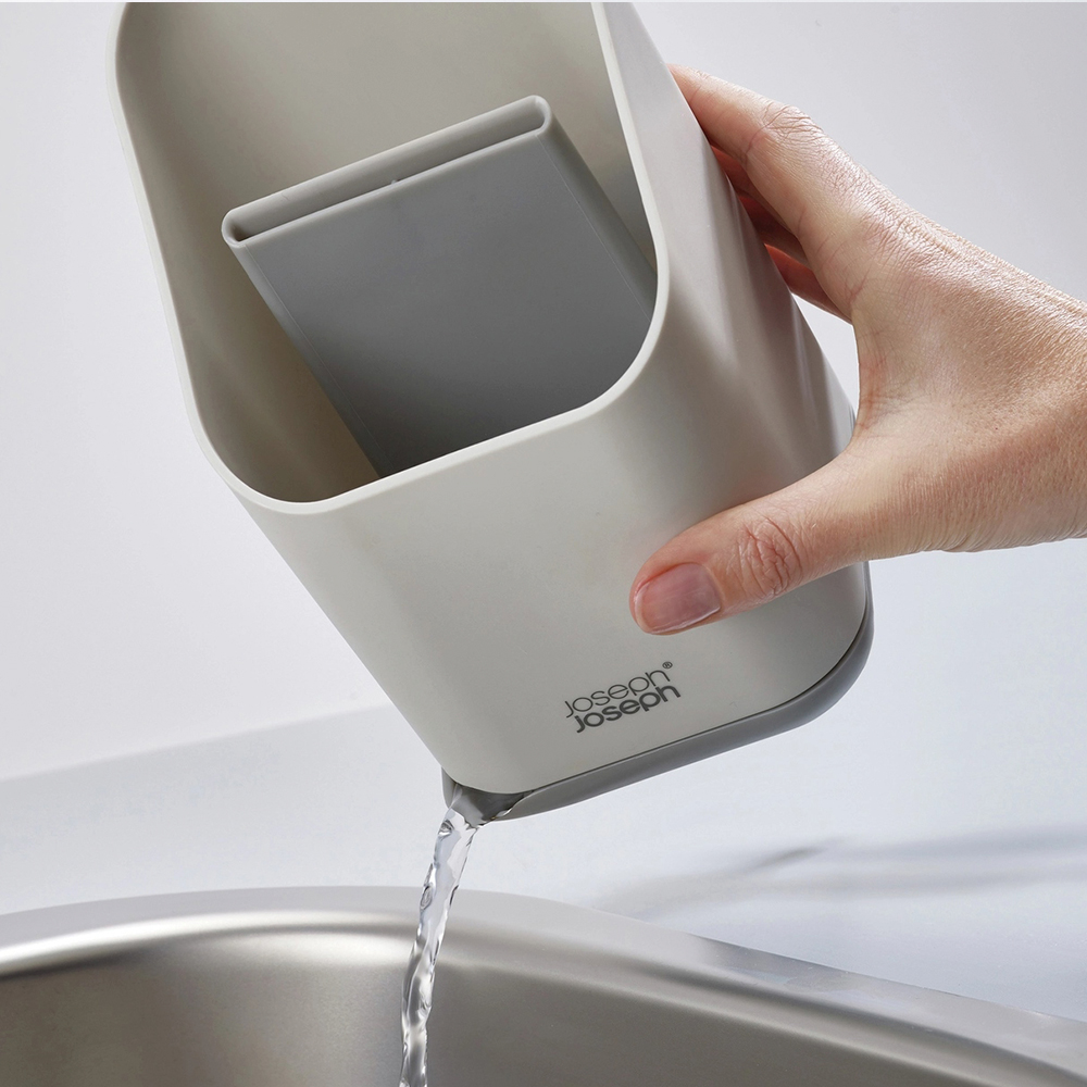 Una mano tiene un contenitore bianco e grigio, versando acqua in un lavandino in acciaio inossidabile. Il contenitore ha un divisore grigio all'interno.