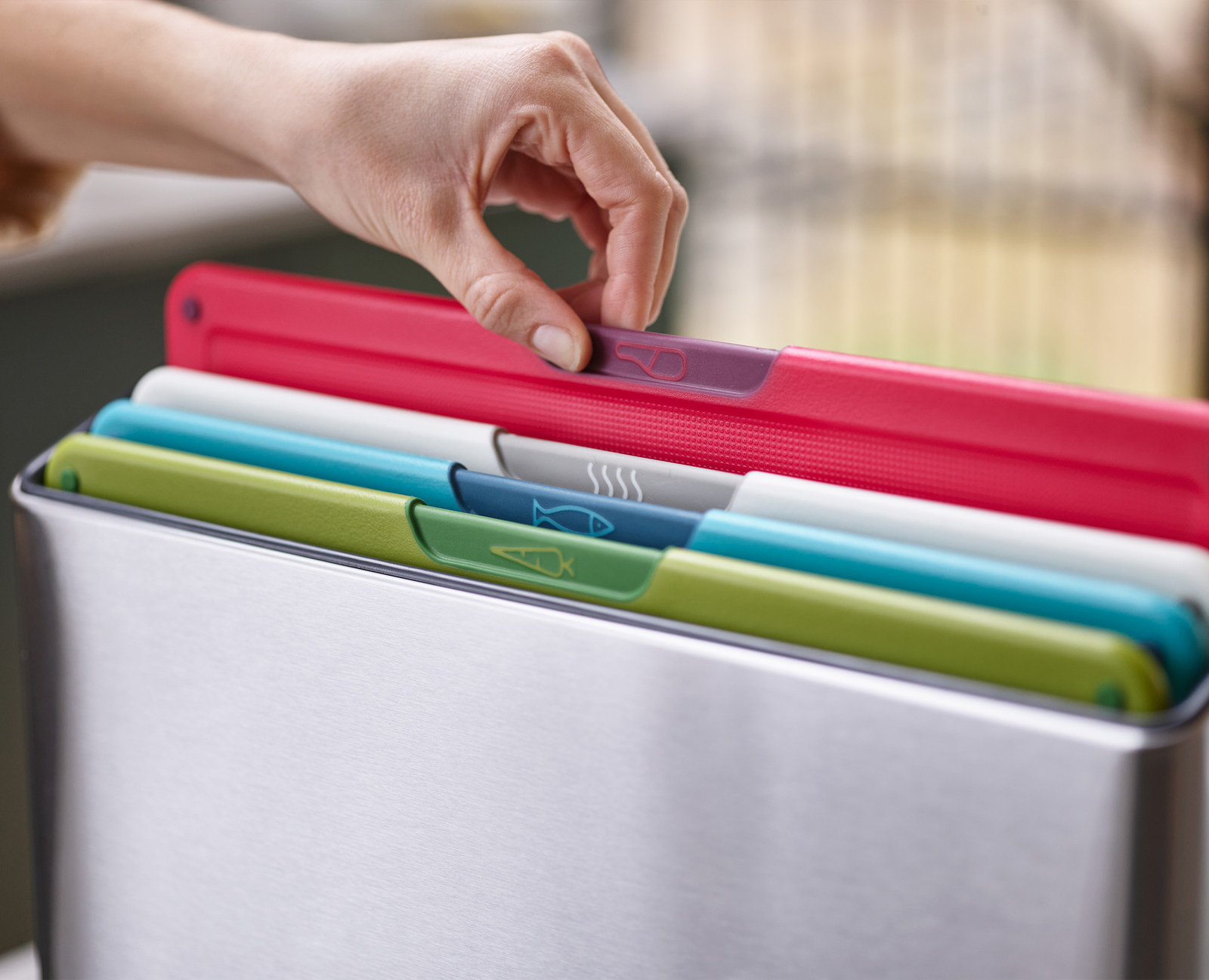 Una mano selecciona una tabla de cortar. Las tablas son de varios colores con iconos de comida. Están en un recipiente de acero inoxidable.