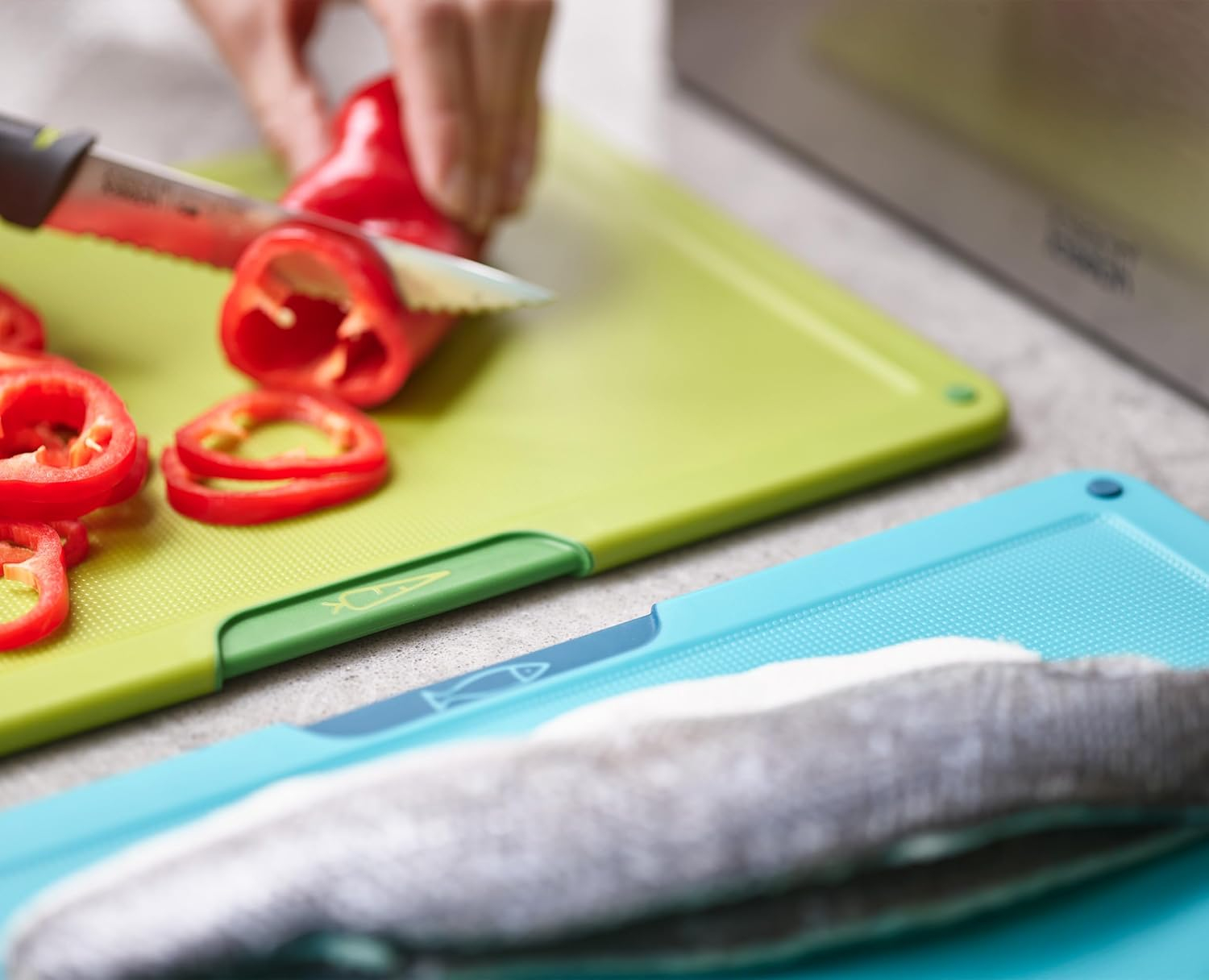 Una persona corta un pimiento rojo en una tabla de cortar verde. Los pimientos en rodajas están en primer plano, con una tabla azul debajo.