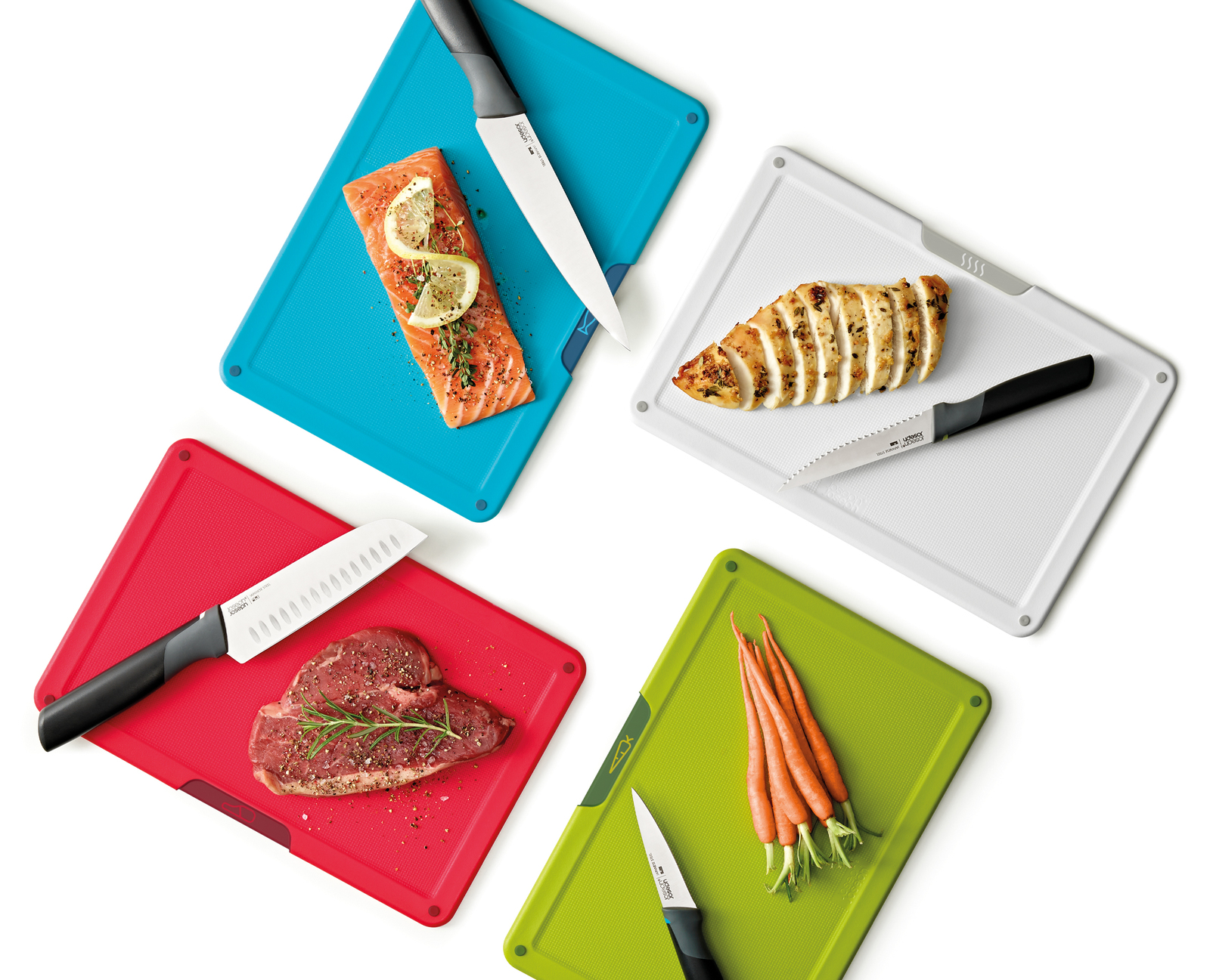 Cuatro tablas de cortar coloridas con comida y cuchillos: salmón, pollo, bistec y zanahorias. Fondo blanco.