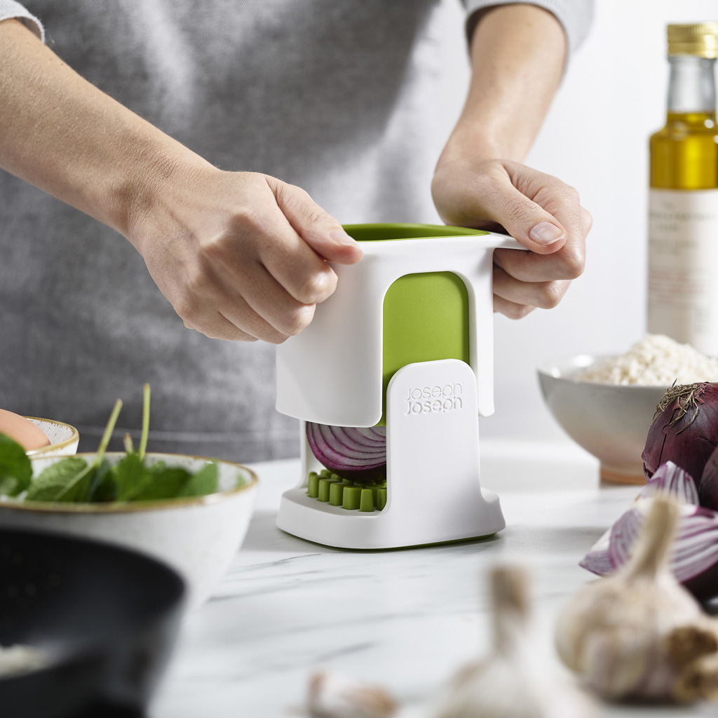 Manos sostienen un picador de cebolla blanco y verde con una cebolla roja dentro. Otros ingredientes, como ajo y aceite, también están en la mesa.