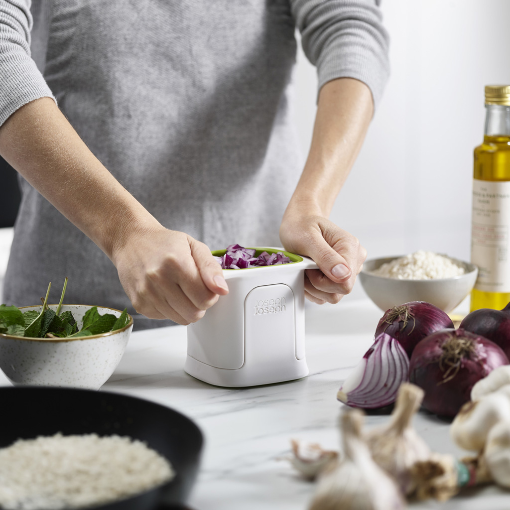 Una persona usa un picador blanco y verde lleno de cebollas rojas. Otros ingredientes como ajo, aceite y arroz están en la mesa.