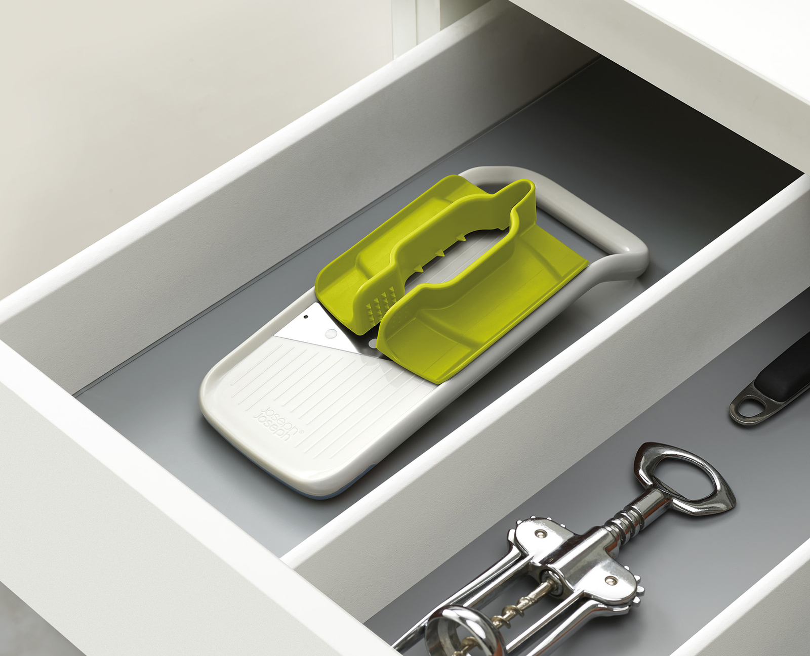 Un cassetto della cucina contiene una grattugia con manico verde, un cavatappi e altri utensili da cucina. Il cassetto è bianco.