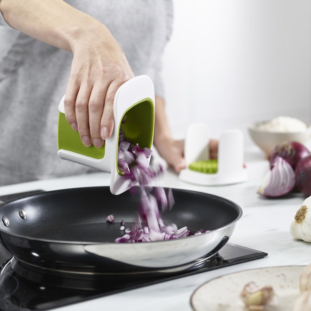 Una persona vierte cebollas rojas cortadas en cubitos de un picador blanco y verde en una sartén negra. Otras herramientas de cocina son visibles.