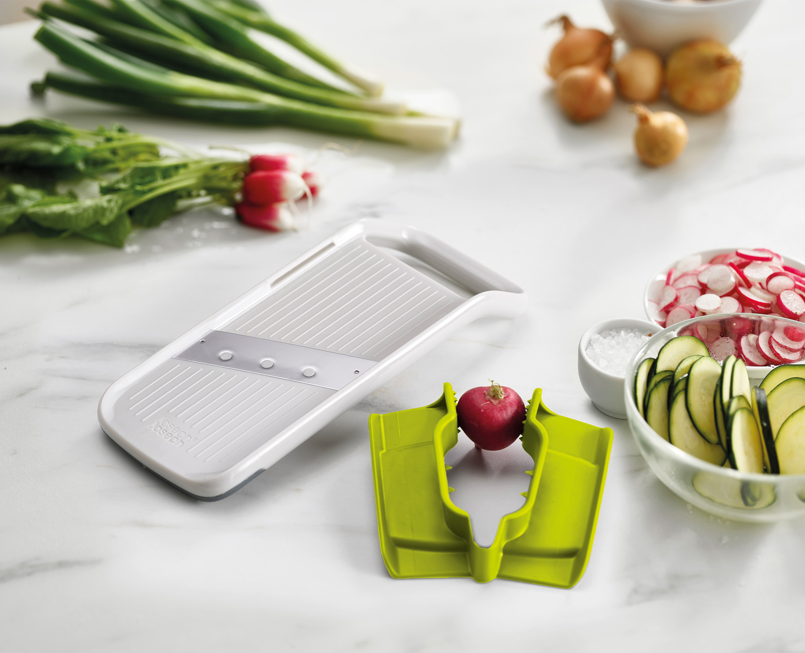 L'immagine mostra un affettatrice per verdure con ravanelli, zucchine a fette, cipolle e cipolline. La scena è su una superficie di marmo bianco.