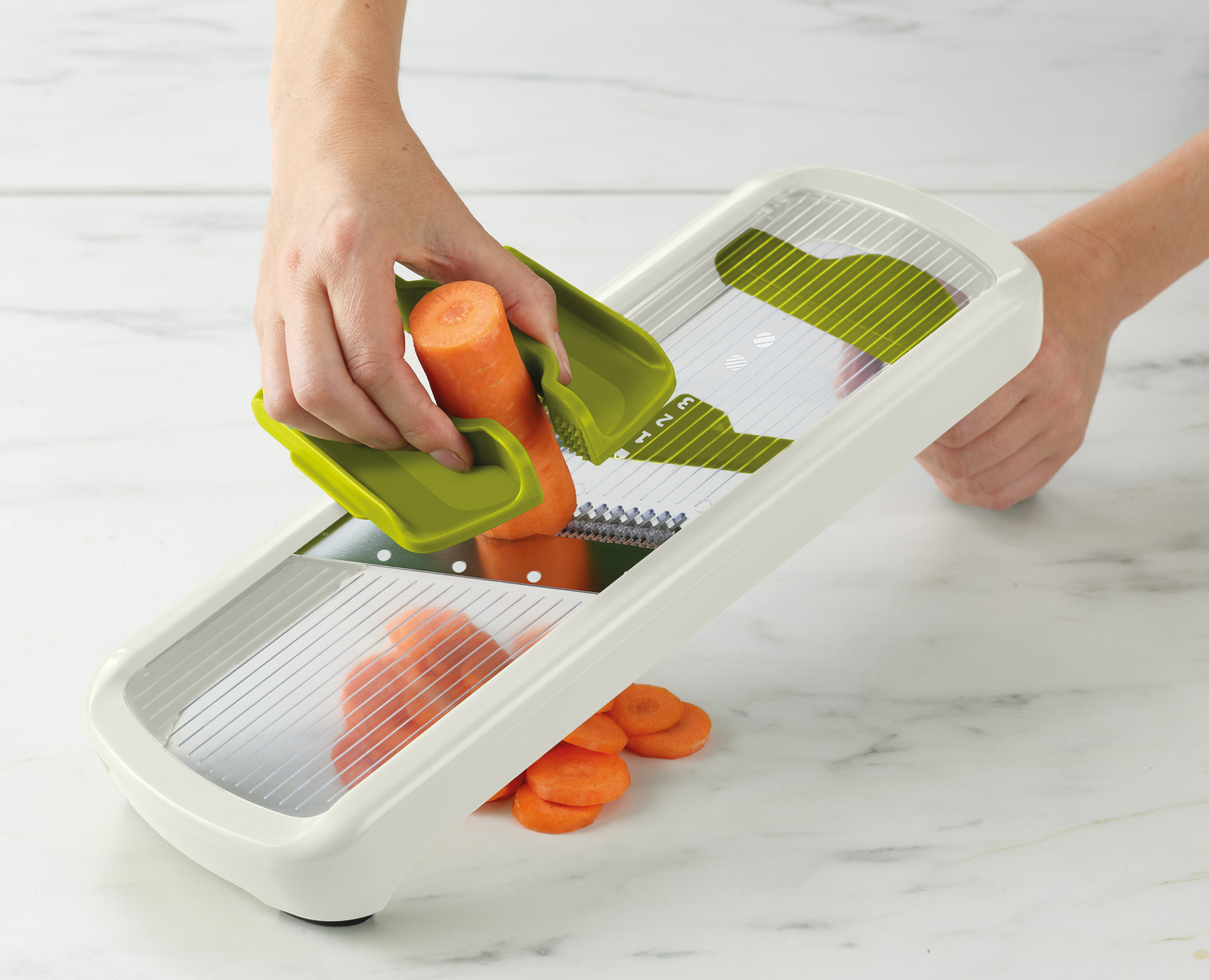 Una persona sta affettando una carota con un affettatrice bianca e verde, con alcune fette già fatte, su una superficie di marmo.