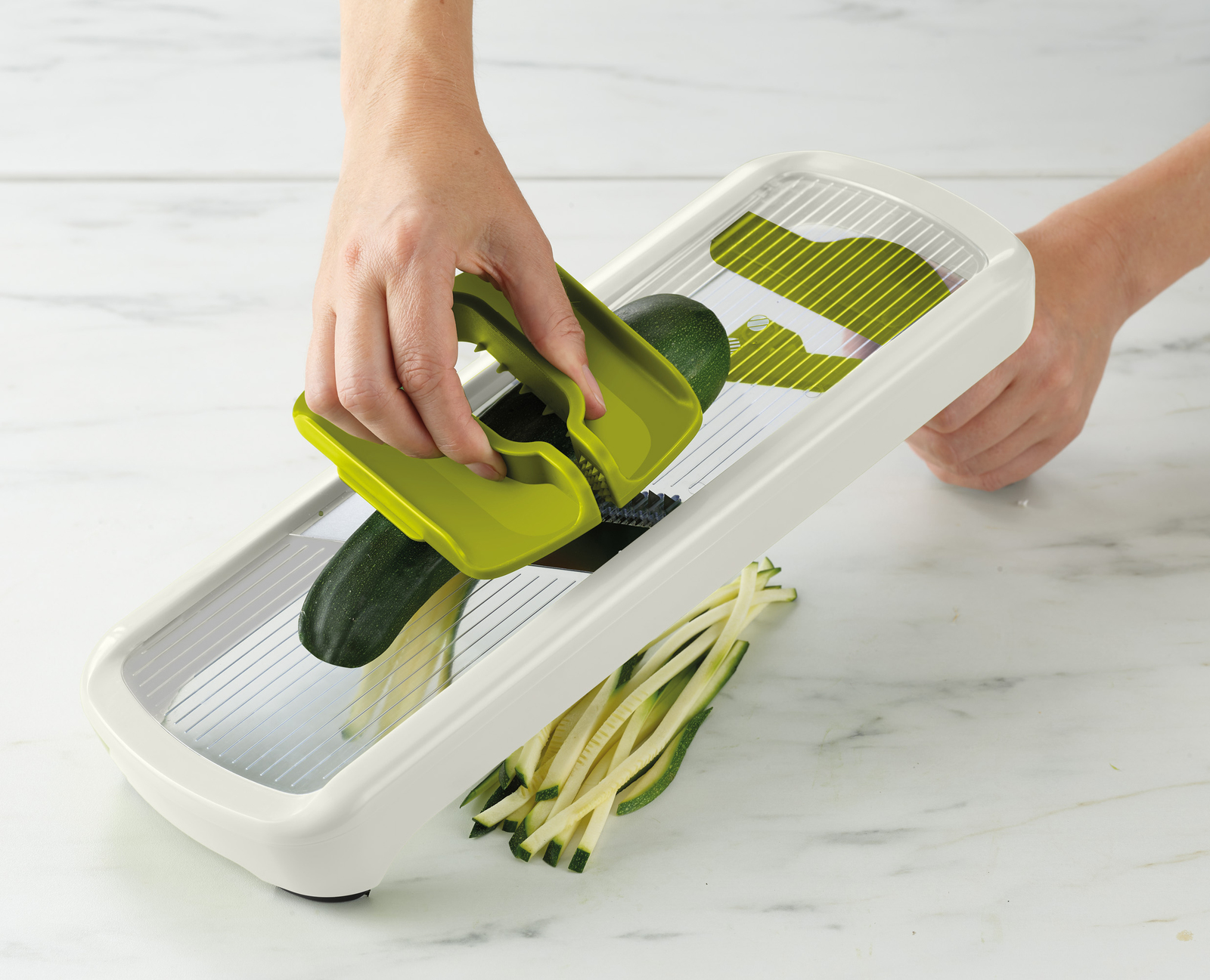 Una persona sta usando un'affettatrice verde per tagliare una zucchina. La zucchina a fette è in primo piano. L'affettatrice è bianca.