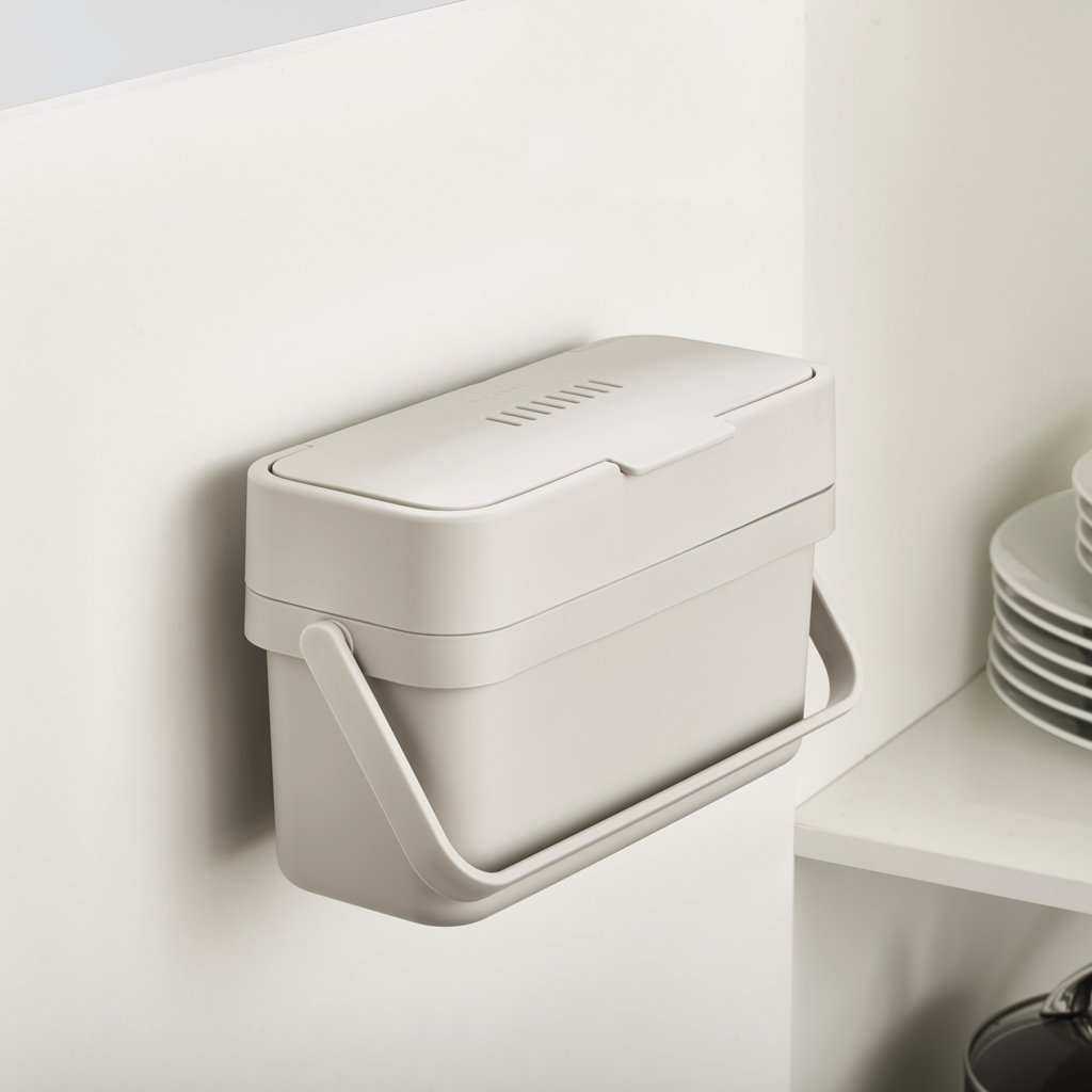 Un cubo de compostaje beige cuelga de una pared blanca. El cubo tiene un asa y una tapa con ranuras. Se ven platos en el fondo.