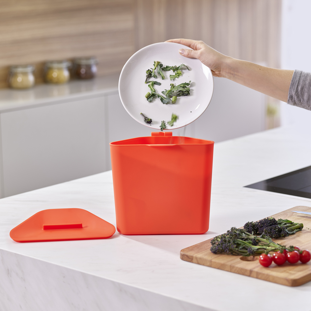 Una mano versa gli scarti di verdura da un piatto in un bidone del compost arancione. Broccoli e pomodori sono su un tagliere.
