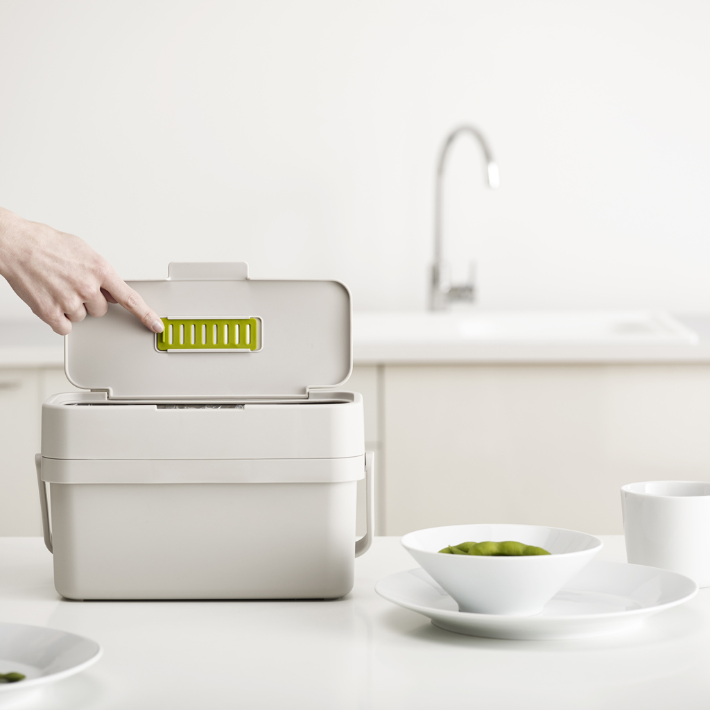 Una mano abre la tapa de un cubo de compostaje beige con una ventilación verde. Un cuenco de judías verdes, un plato y una taza están sobre una mesa blanca.