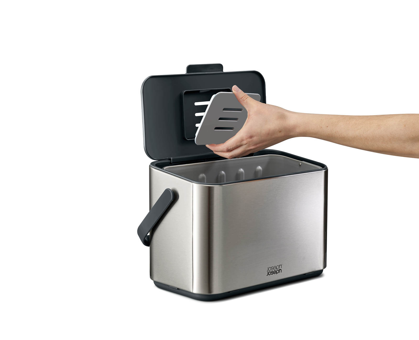 Una mano coloca un utensilio gris en un cubo de acero inoxidable abierto. El cubo tiene tapa y asa negras, y el fondo es blanco.