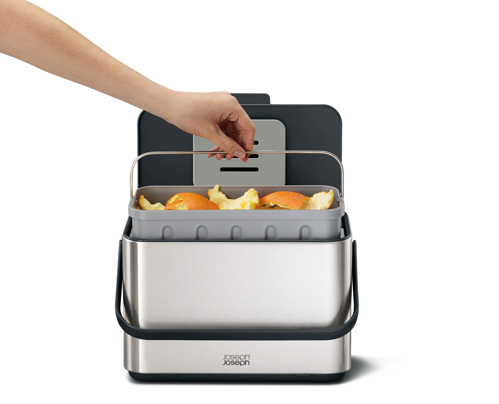 Una mano coloca una cáscara de naranja en un contenedor de compostaje gris. El contenedor es de acero inoxidable, con asa y tapa gris oscuro.
