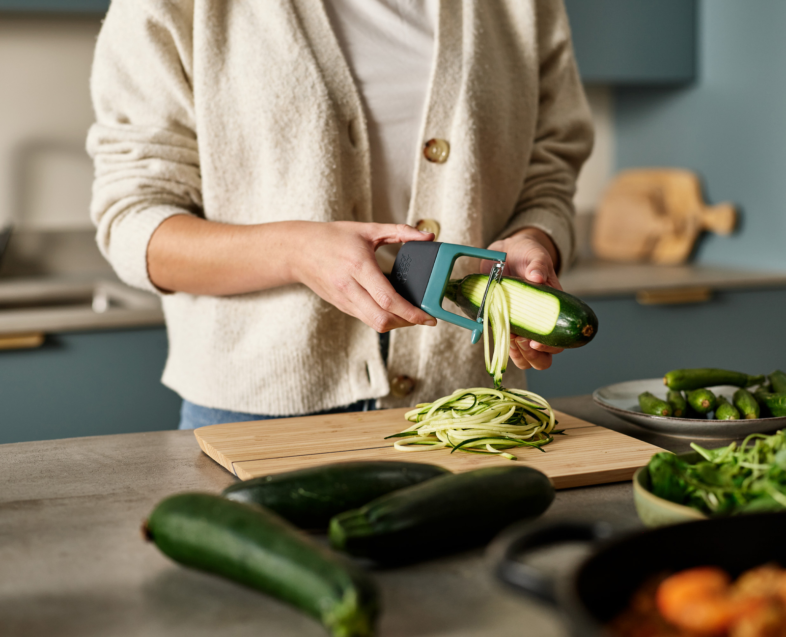 Una persona está espiralizando calabacín sobre una tabla de cortar. Otros calabacines e ingredientes están en la encimera. Ambiente de cocina.