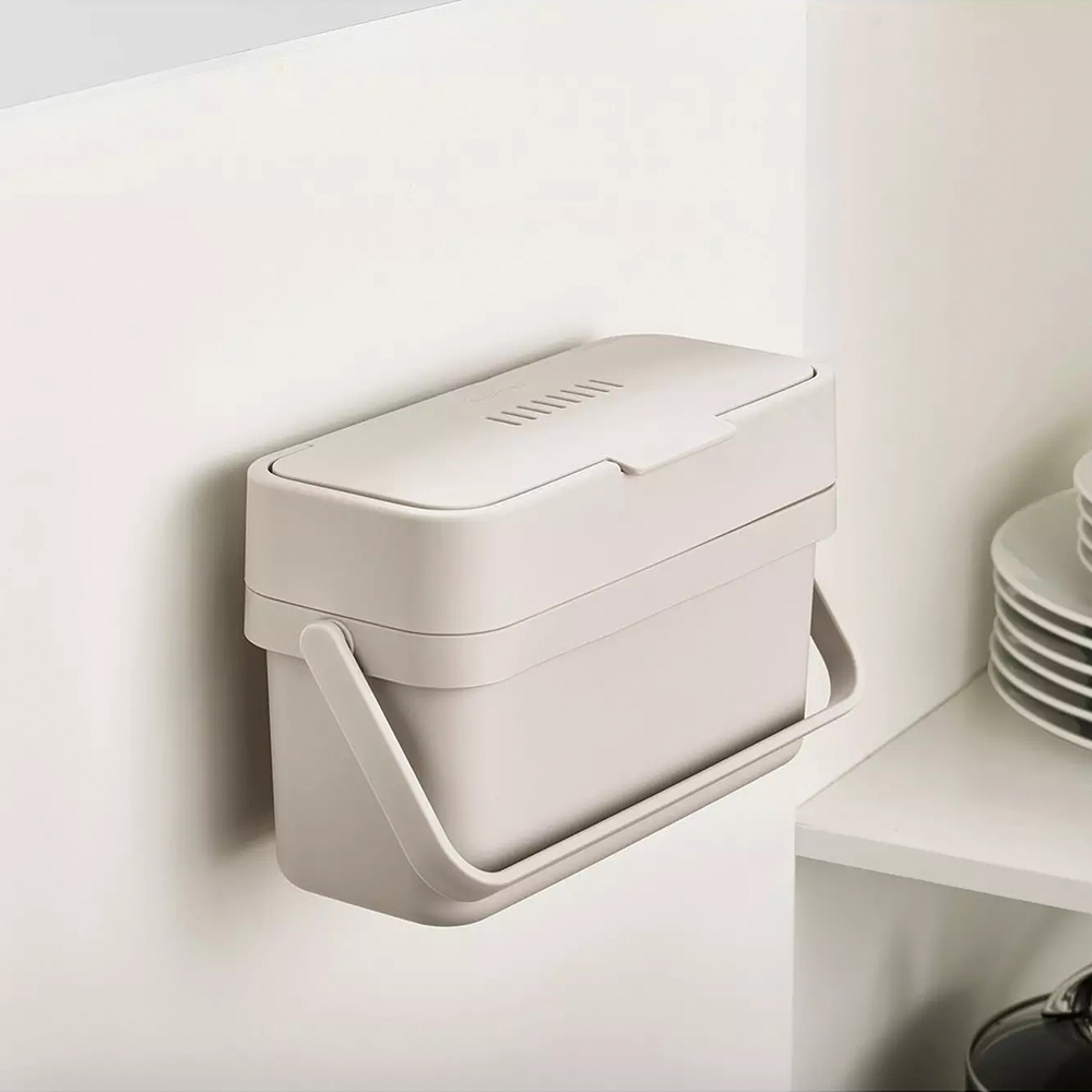Un cubo de compostaje beige y rectangular está montado en una pared blanca. Tiene un asa y una tapa con ranuras de ventilación. Estantes blancos al fondo.