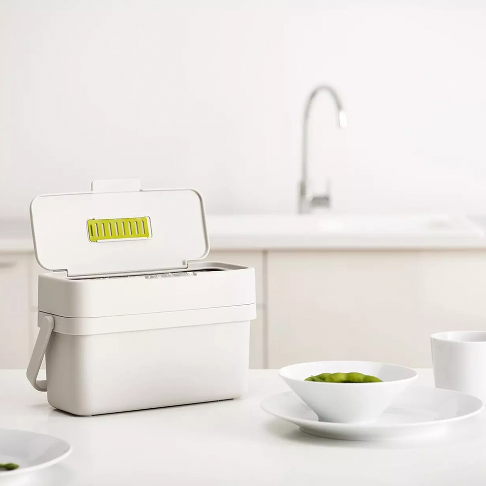 Un contenedor de compostaje blanco con tapa abierta, un cuenco de judías verdes y una taza blanca sobre una mesa blanca. Cocina al fondo.