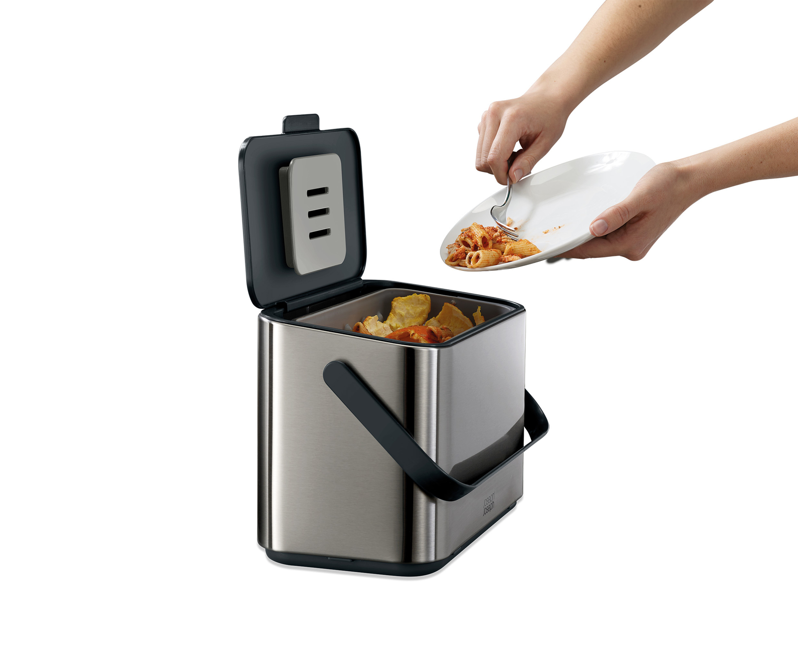 Una persona raspa pasta de un plato en un cubo de compostaje de acero inoxidable. El cubo está abierto y se ven restos de comida dentro.