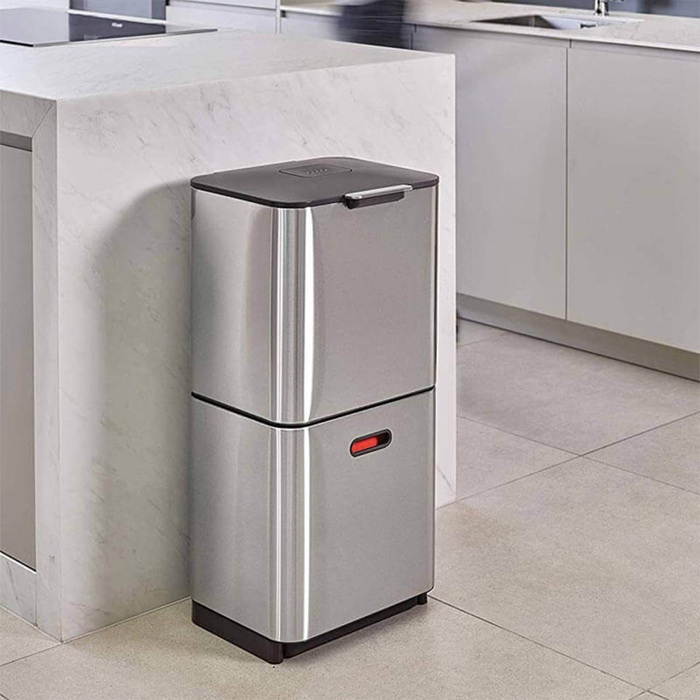 Un bidone della spazzatura in acciaio inossidabile a due sezioni con coperchio nero e spia rossa, su sfondo cucina moderna.