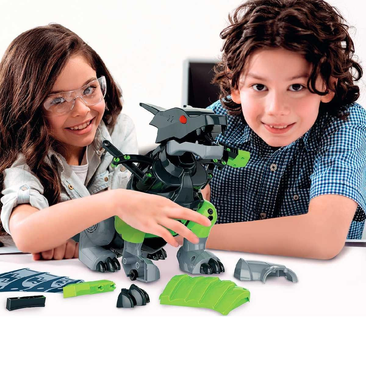 Dos niños ensamblan un dinosaurio robot verde y gris. Las piezas están dispersas sobre una mesa blanca. Están sonriendo a la cámara.