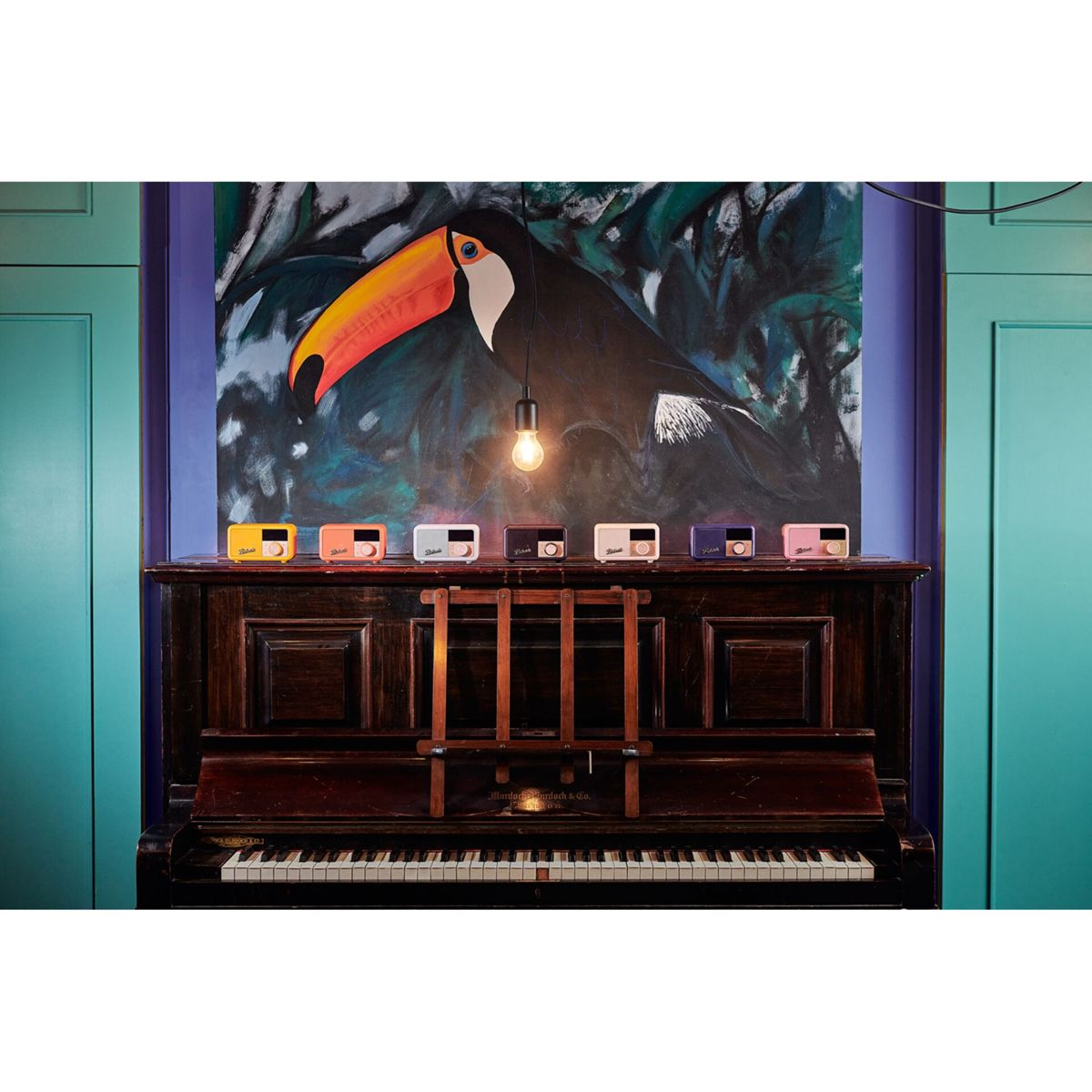 Fortepian z kolorowymi radiami na górze. Obraz tukan wisi za fortepianem, nad nim żarówka. Ściana jest turkusowa i fioletowa.