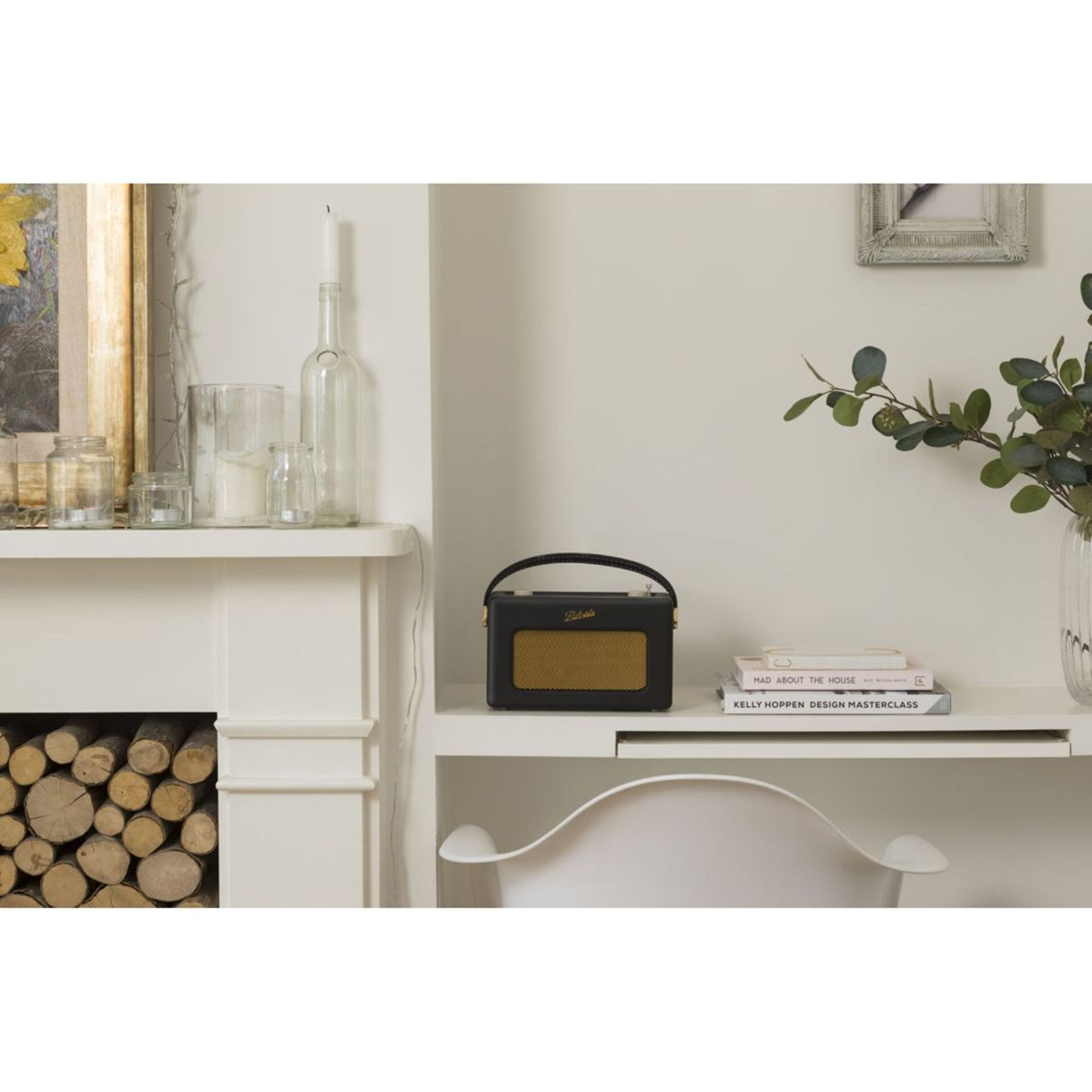Czarno-złote radio stoi na białej półce z książkami. Przed nim białe krzesło. Drewno opałowe ułożone w kominku.