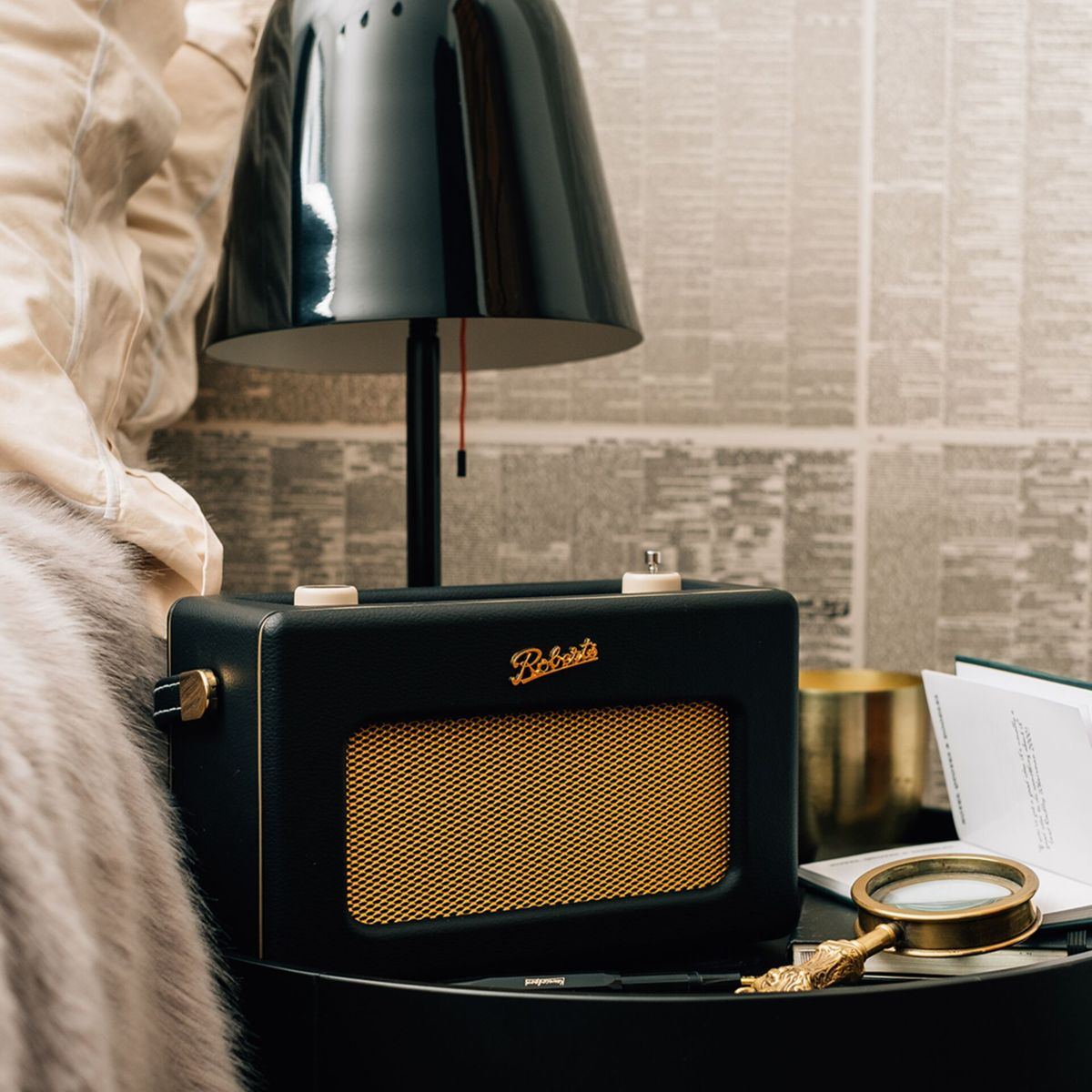 Czarny radio z złotym głośnikiem na czarnym stole. Na stole jest też lampa i lupa. Po lewej stronie jest łóżko.