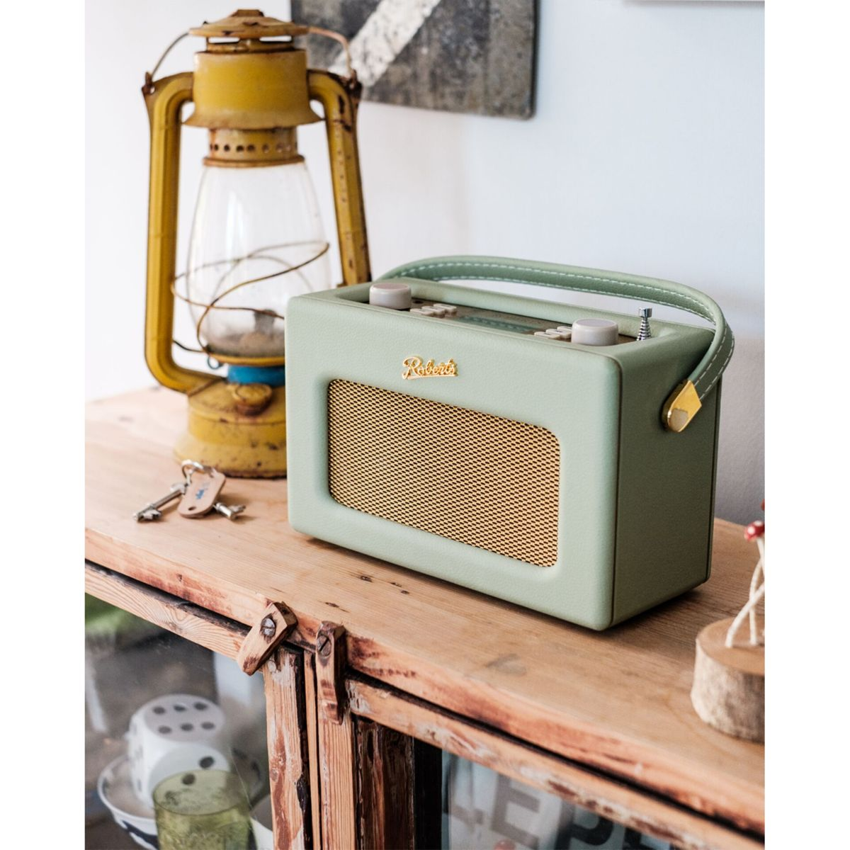 Miętowe radio ze złotym głośnikiem i uchwytem stoi na drewnie, obok żółtej latarni i kluczy. Tło to sztuka abstrakcyjna.