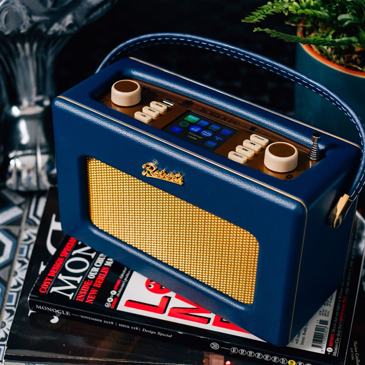 Niebieskie radio Roberts ze złotym przodem z siatki stoi na magazynach. Na górze jest uchwyt. Mała roślina w tle.