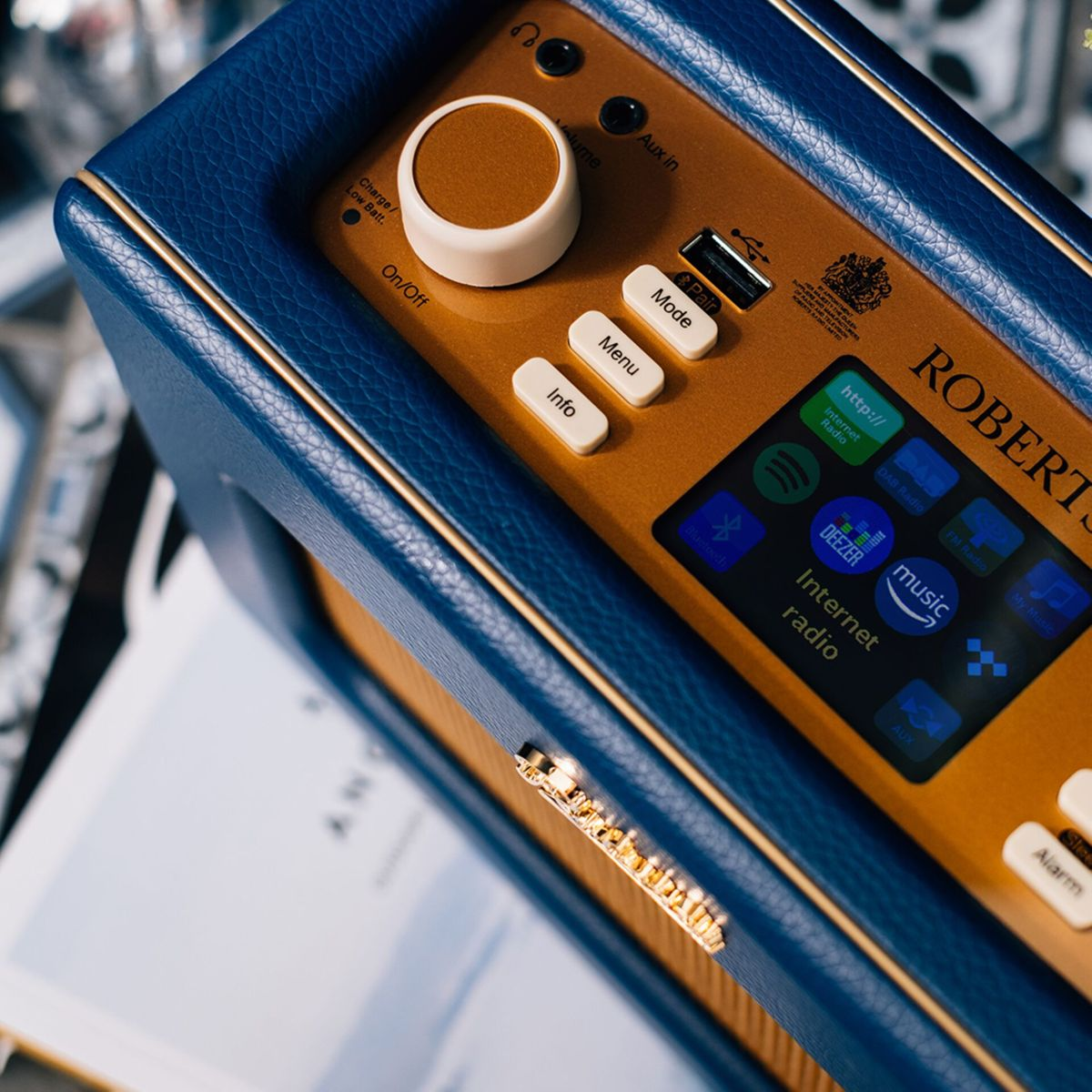 Zbliżenie niebiesko-złotego radia Roberts. Ma przyciski, ekran i pokrętło głośności. Tło jest rozmyte.