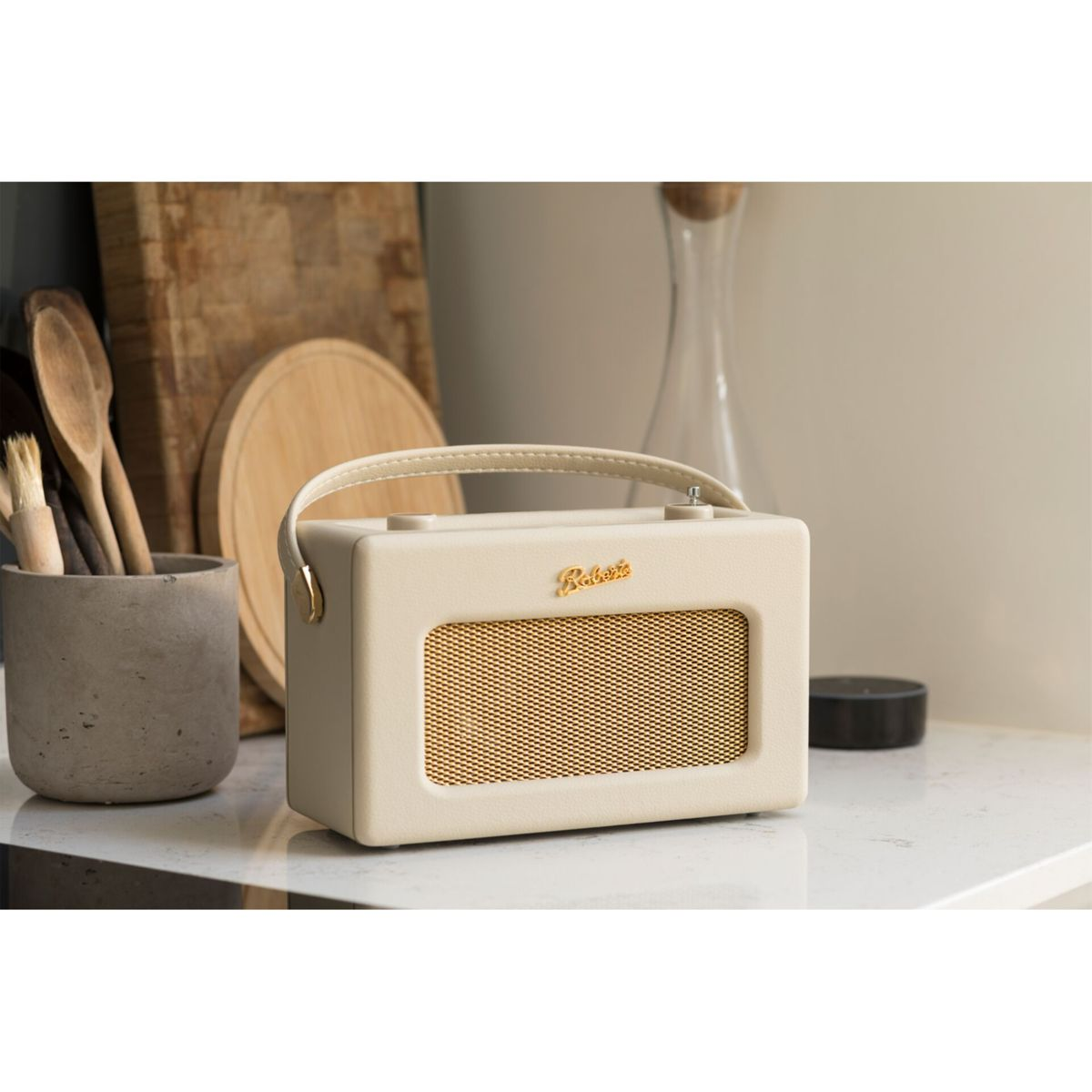 Kremowe radio Roberts stoi na białym blacie. Drewniane przybory kuchenne i deski do krojenia znajdują się w tle.