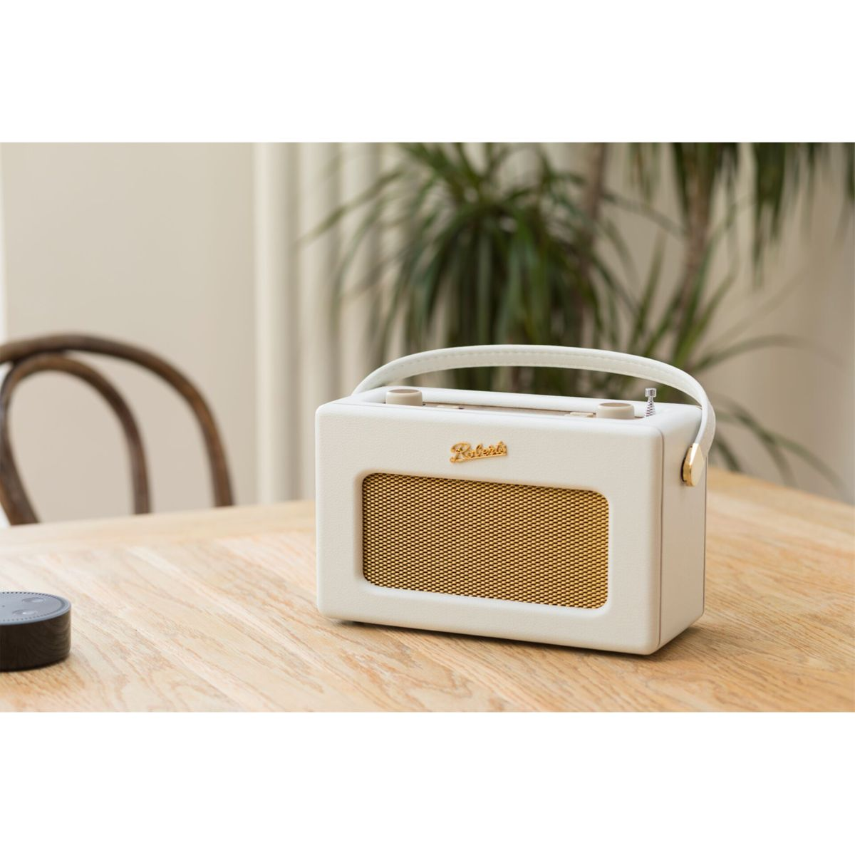 Kremowe radio Roberts stoi na drewnianym stole. Po lewej stronie znajduje się czarny głośnik inteligentny. W tle roślina.
