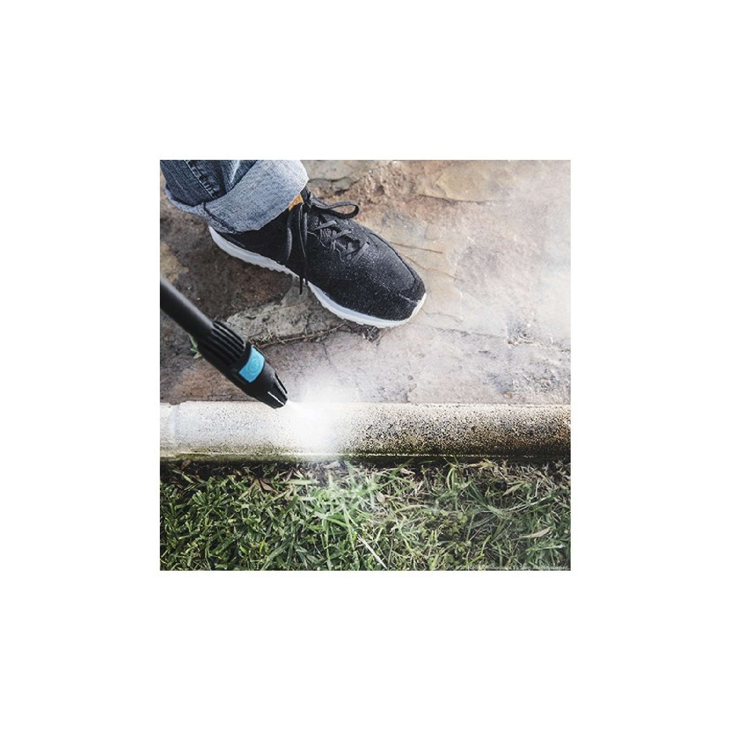 Una persona usa una hidrolavadora en una superficie de hormigón. Se ve un zapato negro. Hay hierba debajo del hormigón.