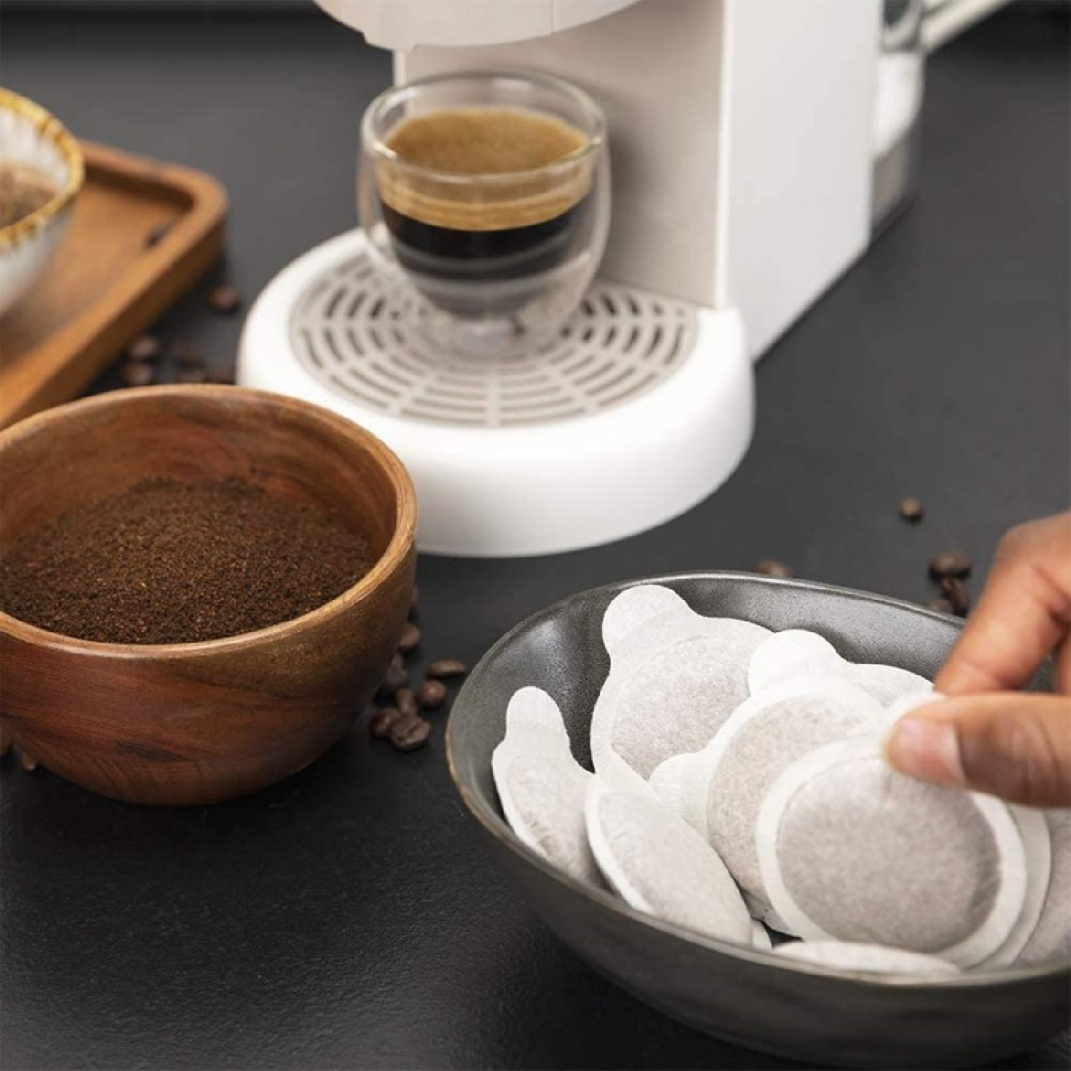 Eine Kaffeemaschine mit einem Glas Kaffee, Kaffeepulver in einer Holzschale und Kaffeepads in einer Keramikschale sind zu sehen.