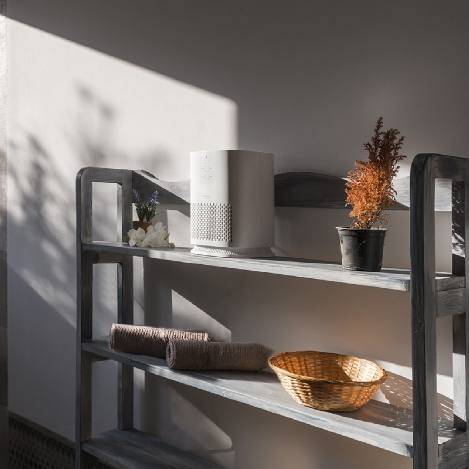 Un purificatore d'aria bianco su una mensola di legno con asciugamani, un cestino e una pianta in vaso. La luce solare e le ombre evidenziano gli oggetti.