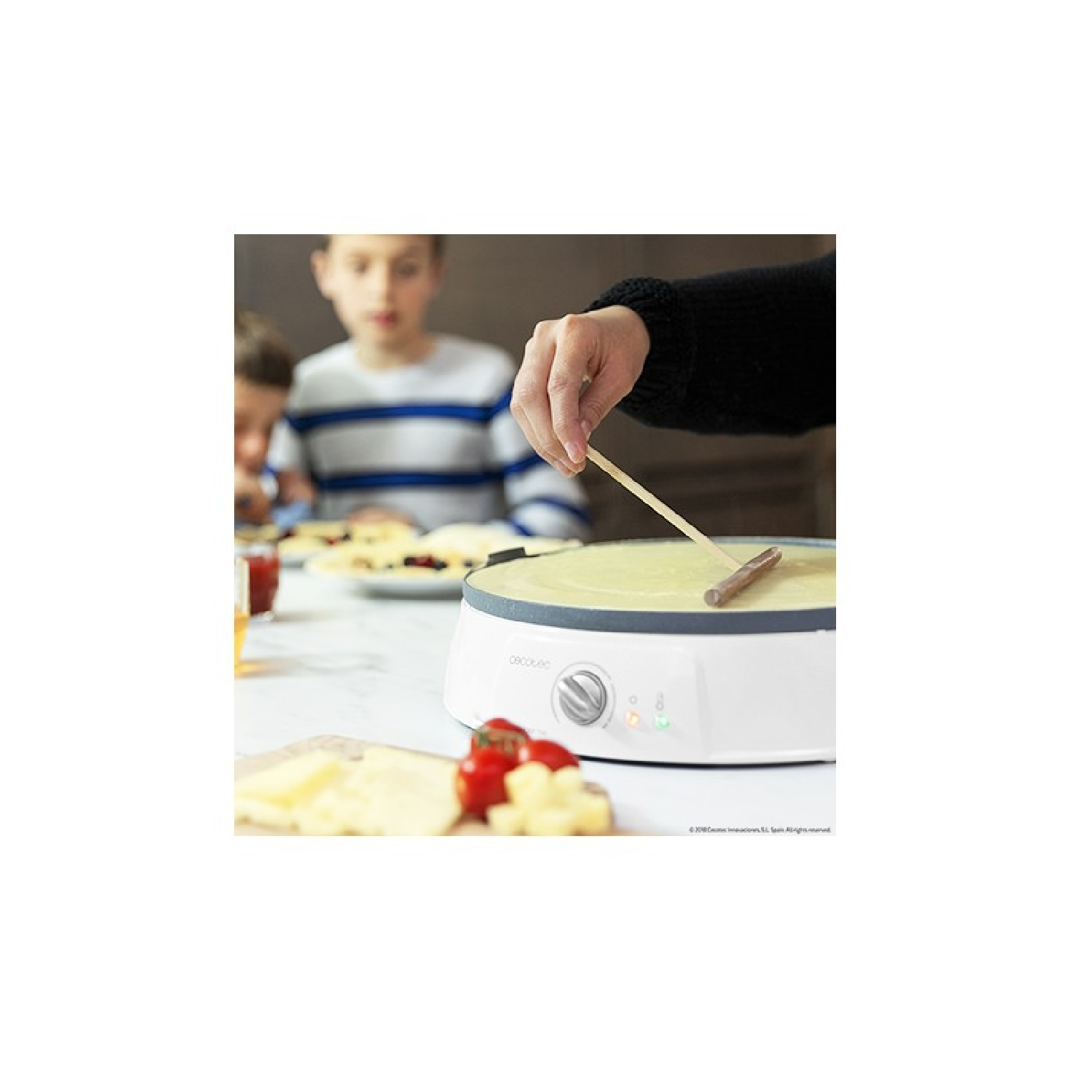 Een persoon gebruikt een houten spatel om beslag op een witte crêpemaker te smeren. Kaas, tomaten en kinderen op de achtergrond.