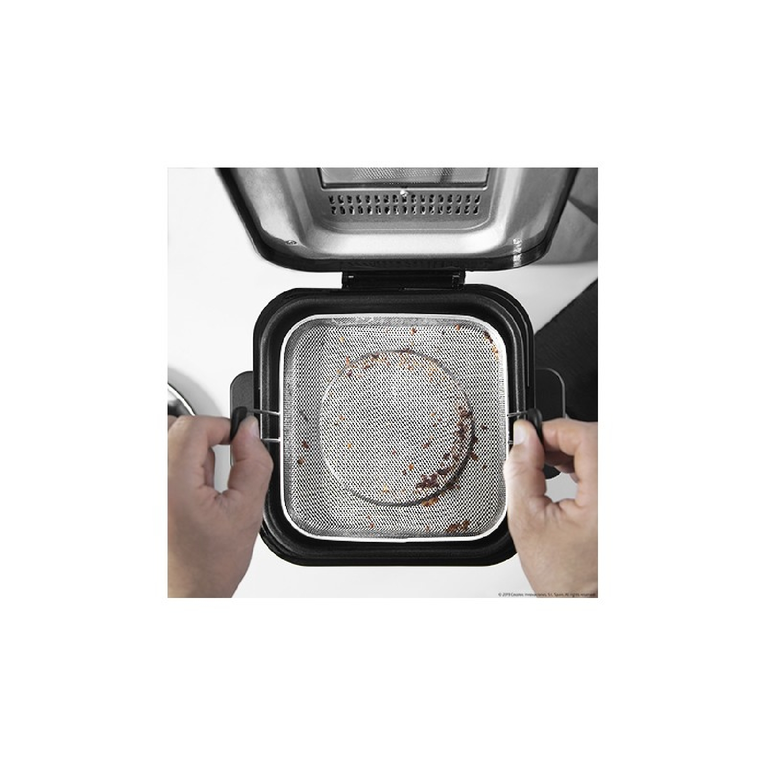 Dłonie trzymają kosz z siatki wewnątrz frytkownicy. Kosz zawiera resztki jedzenia. Frytkownica jest otwarta na białej powierzchni.