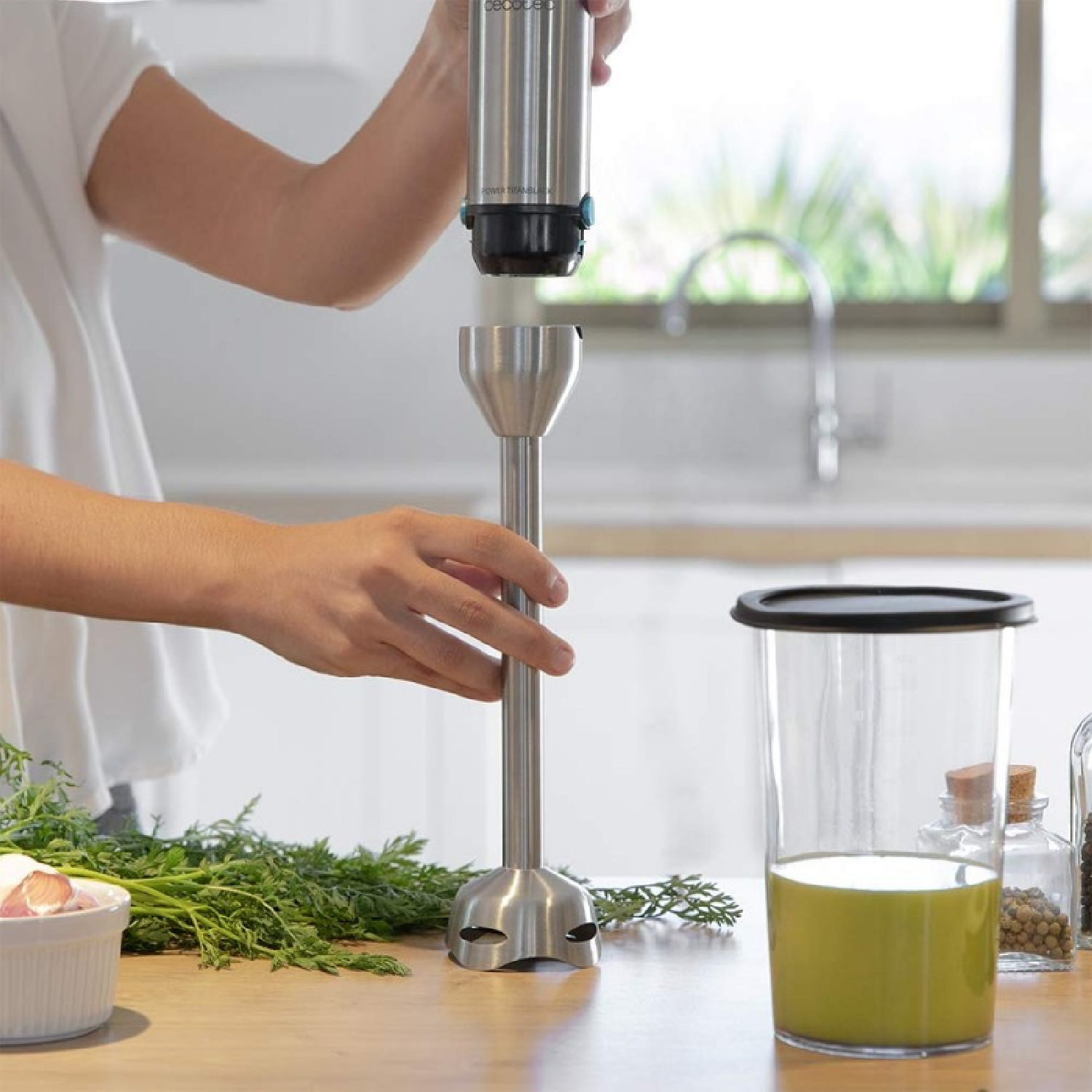 Una persona che assembla un frullatore a immersione. Verdure, una tazza di liquido verde e spezie sono sul tavolo in cucina.