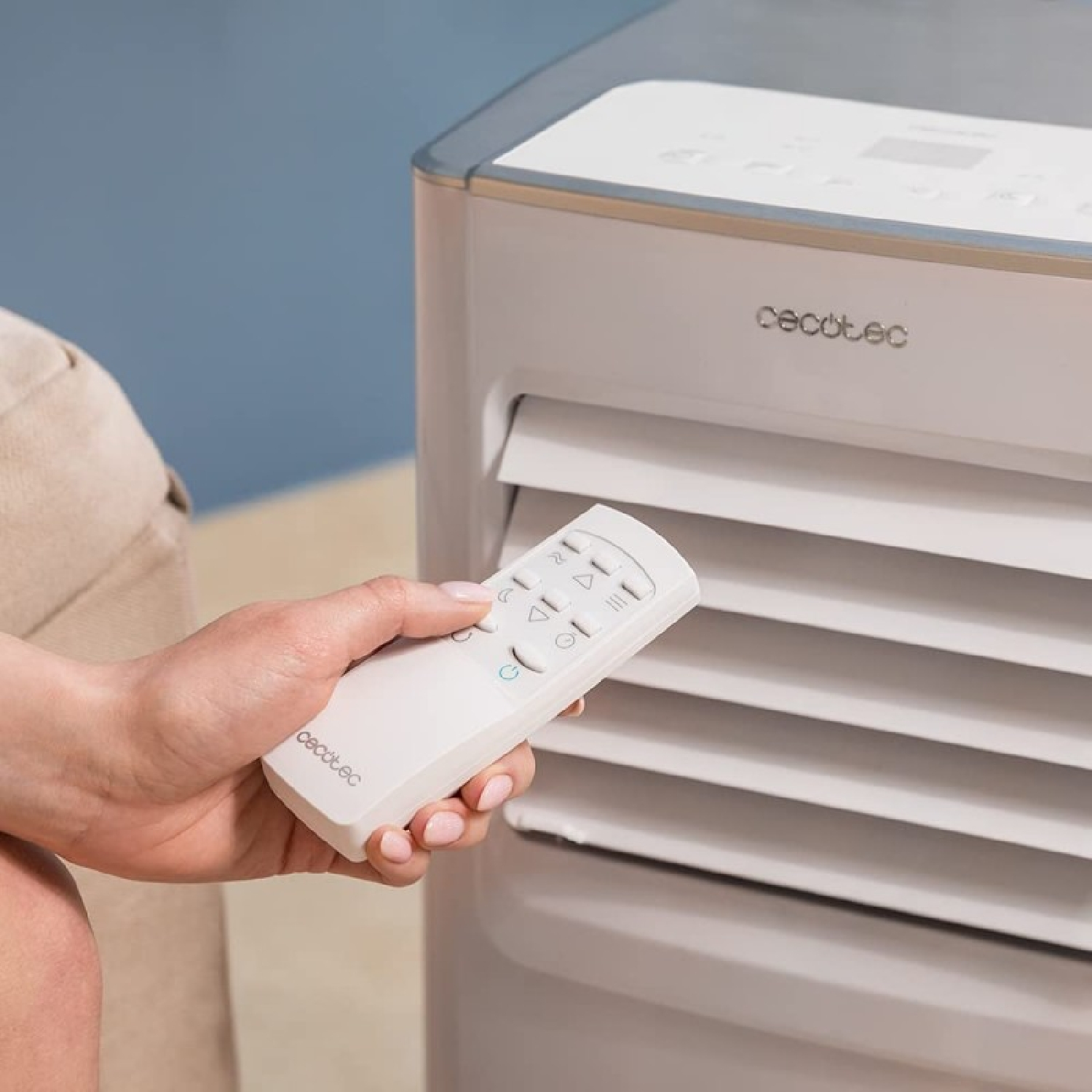 Ręka trzyma biały pilot w pobliżu biało-szarej chłodziarki powietrza. Tło to niebieska ściana.