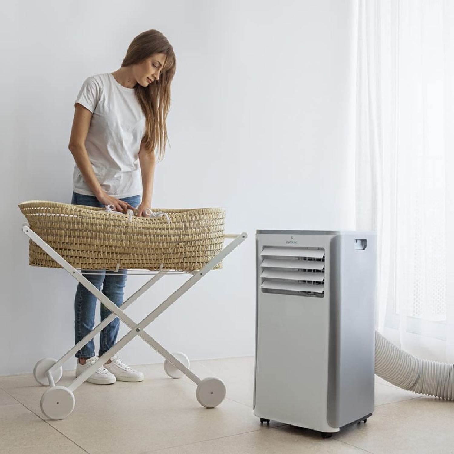 Kobieta stoi blisko łóżeczka dziecięcego, w pobliżu klimatyzator. Pokój jest biały z oknem. Łóżeczko jest tkane.