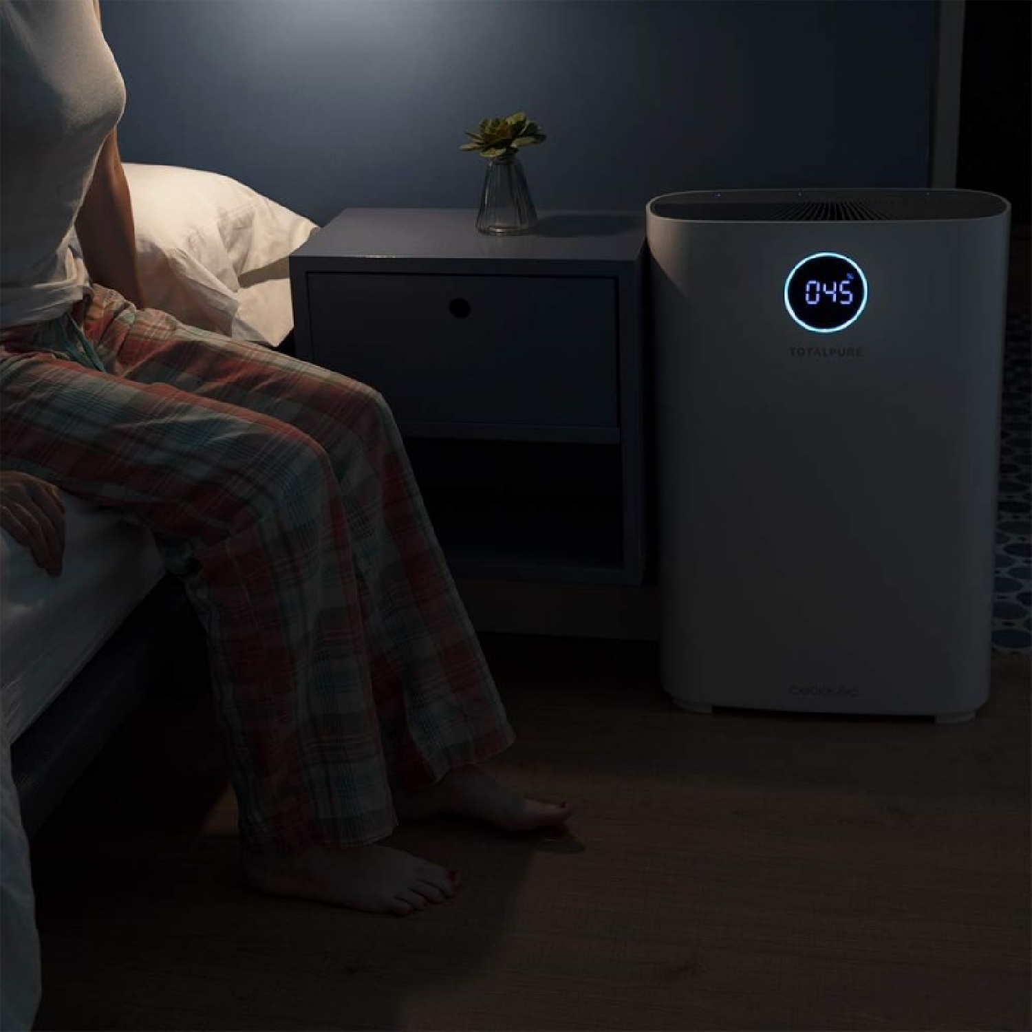 Una persona con pantaloni a quadri è seduta su un letto accanto a un comodino. Un purificatore d'aria con display digitale è sul pavimento.
