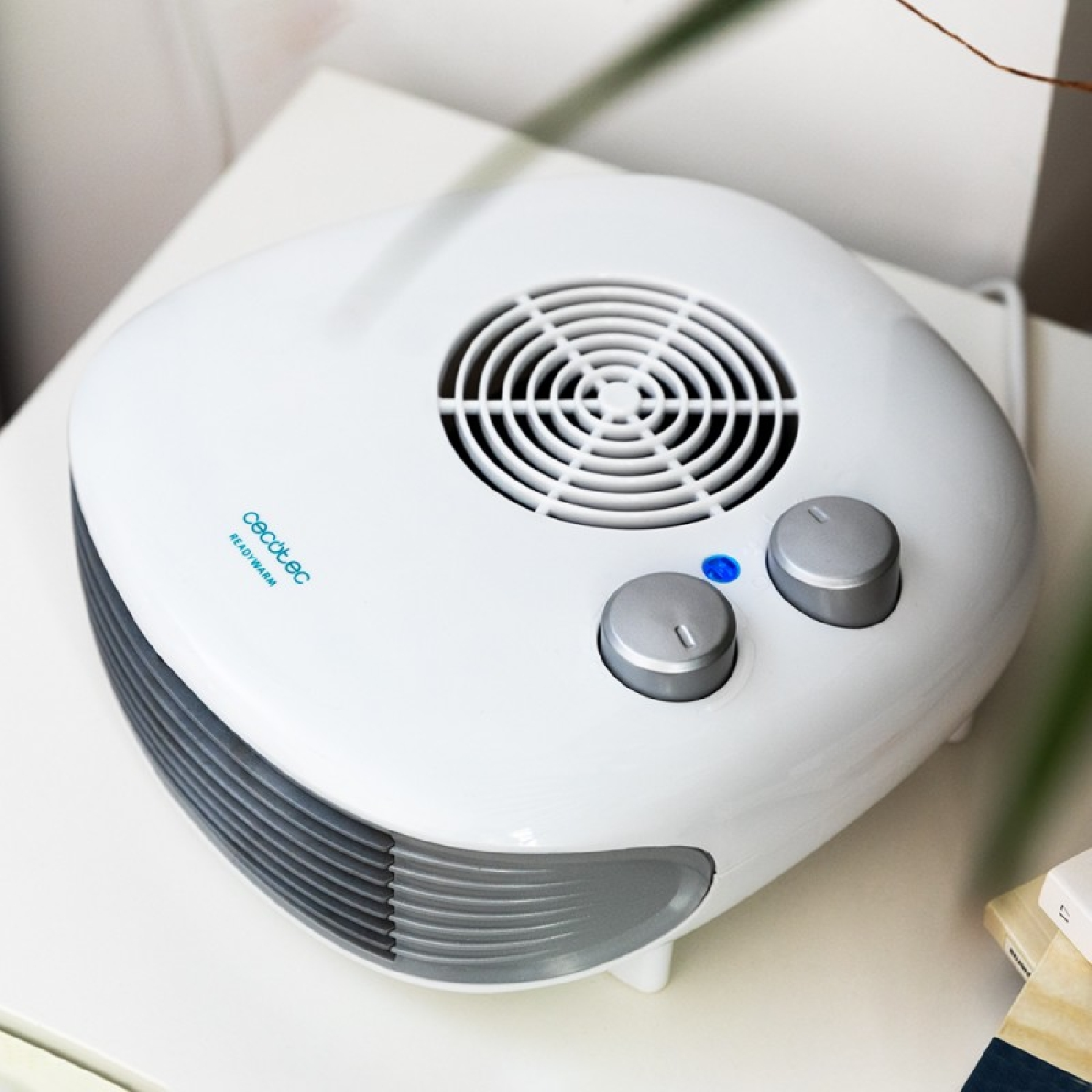 Biały grzejnik elektryczny na białej powierzchni. Ma dwa pokrętła, wentylator i niebieskie światło.