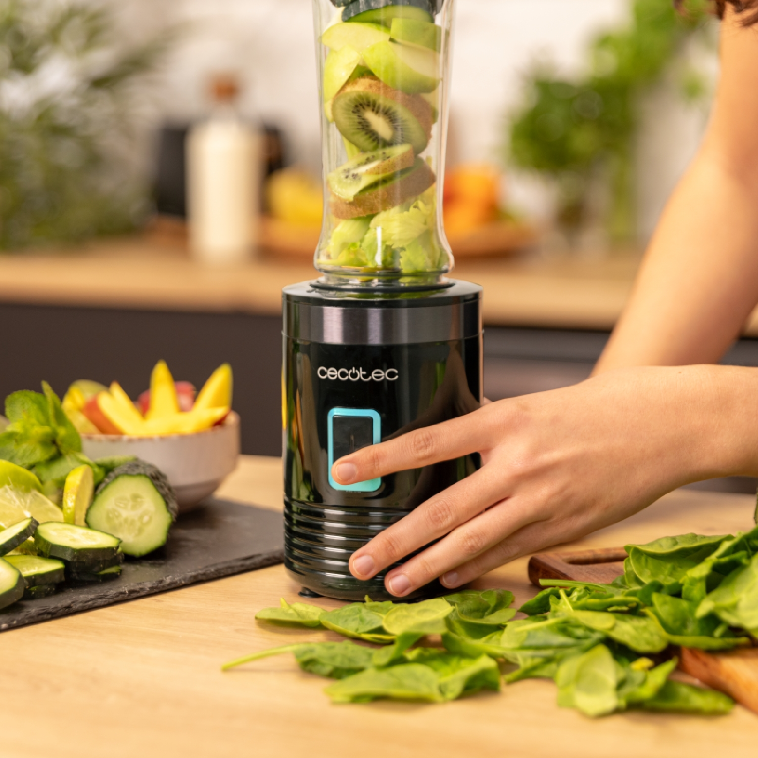 Een blender met fruit en groenten. Een hand drukt op een knop. Spinazie en andere ingrediënten op tafel.