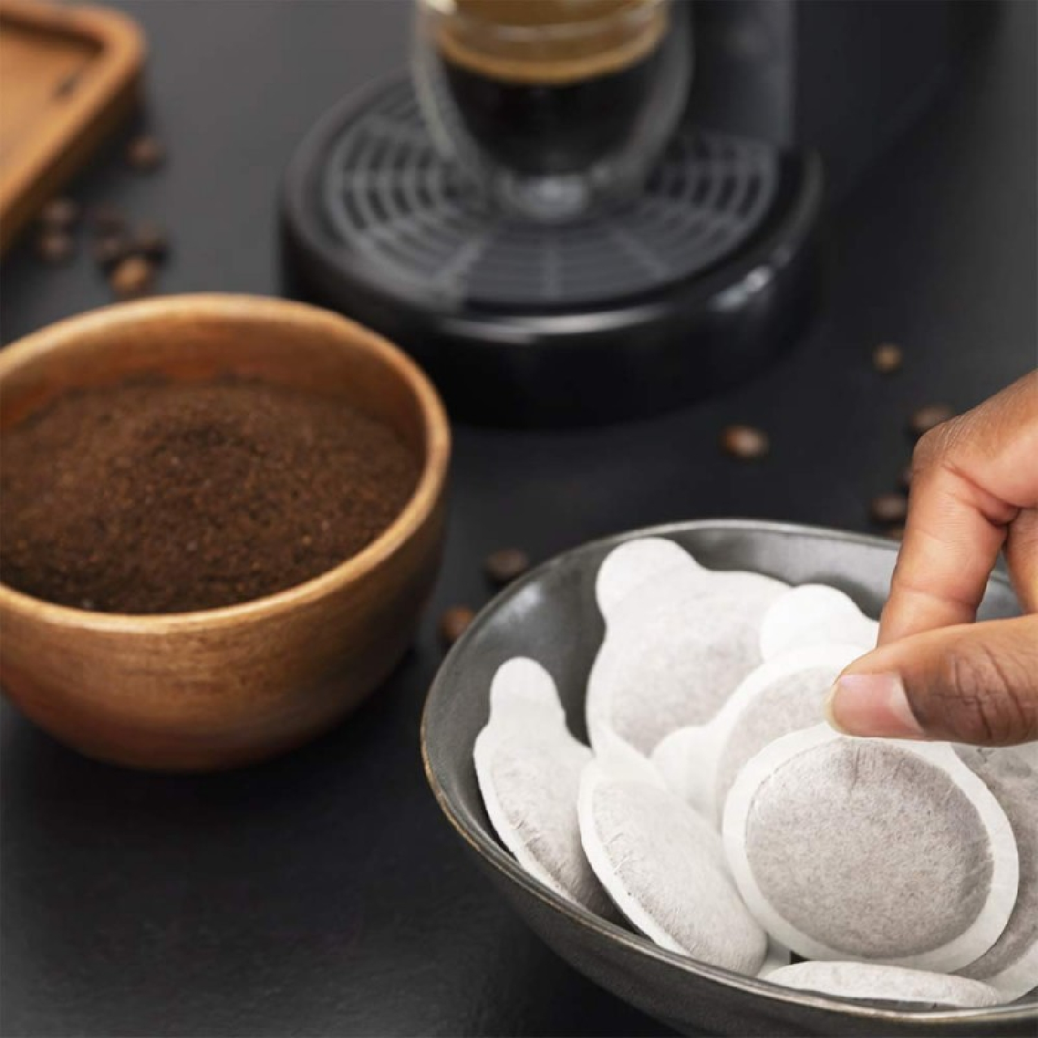 Ręka sięga po kapsułkę kawy z miski. Kawa mielona w drewnianej misce, ekspres do kawy i ziarna kawy są również obecne.